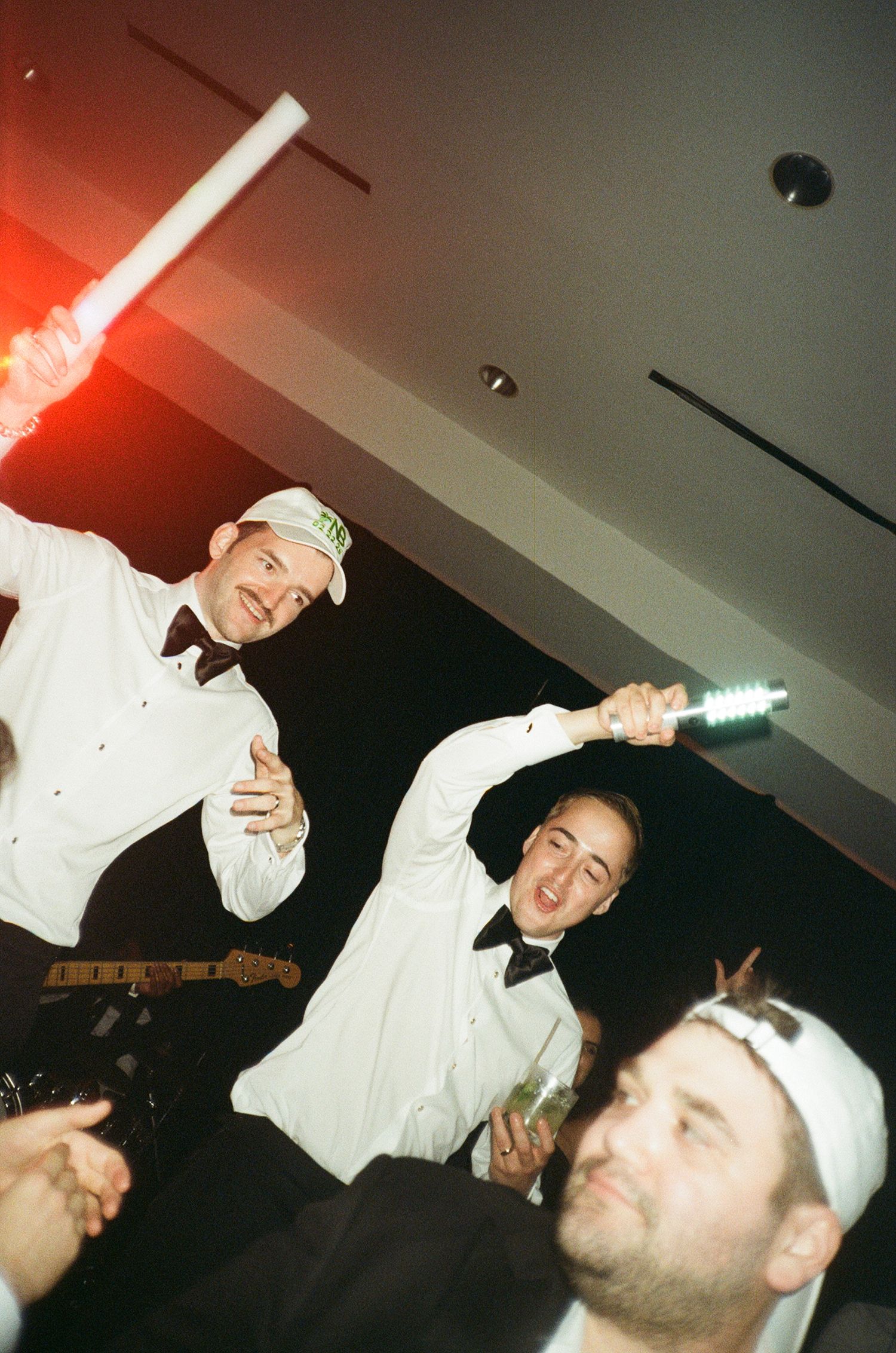 Party guests in white dress shirts celebrating energetically at a dimly lit indoor event with red lighting effects.