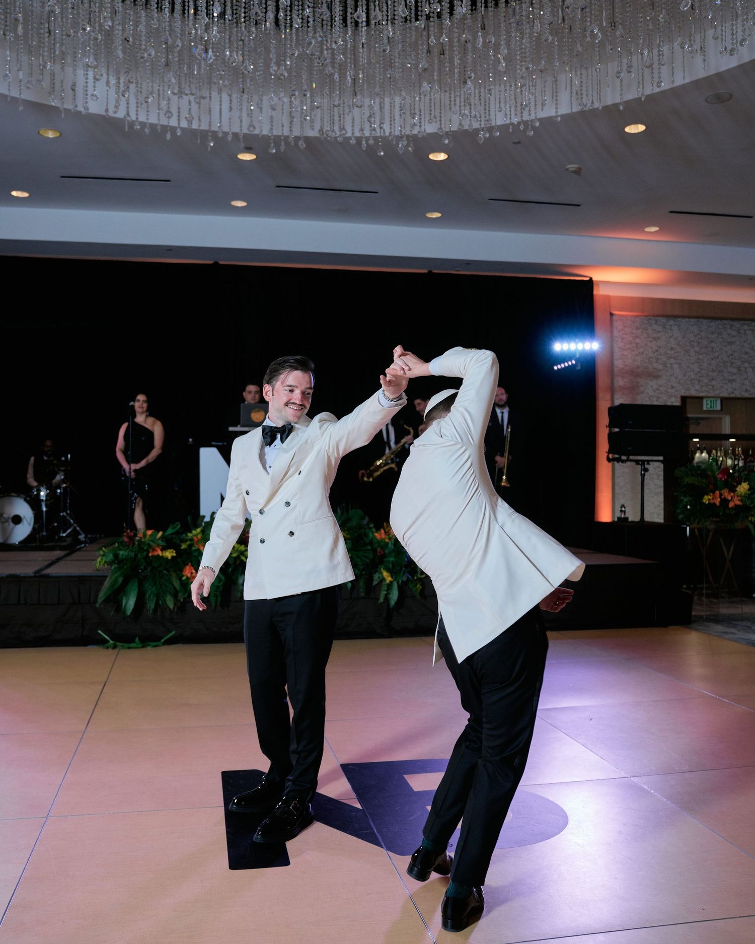 Two people performing an energetic dance move together on an illuminated dance floor beneath a crystal chandelier.