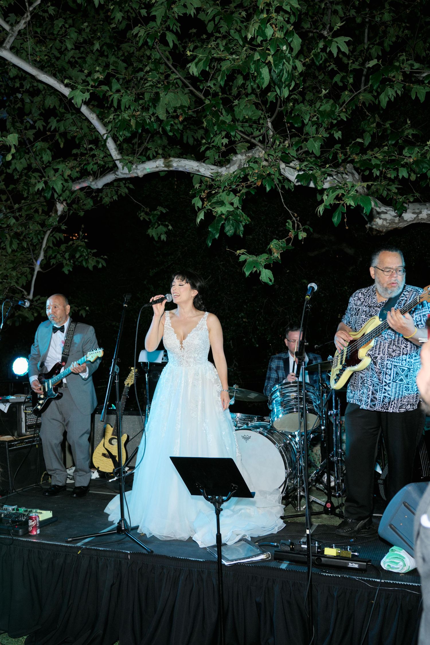 Wedding reception band performs on stage at an outdoor evening event with trees in the background.