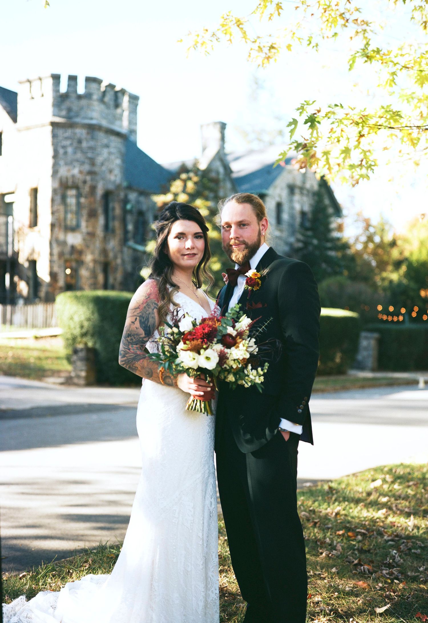 Newlyweds pose together in front of a stone building with an autumn backdrop and floral bouquet.