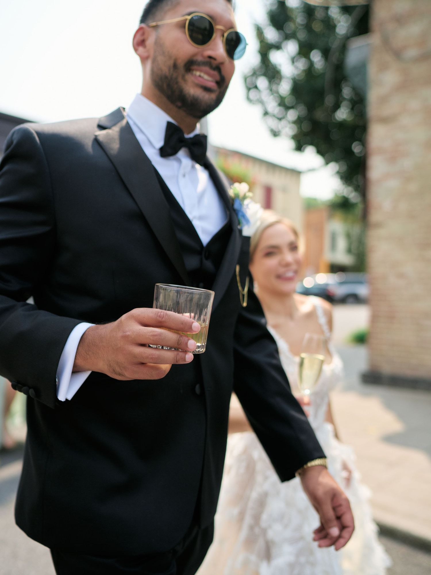 Elegant couple at a wedding celebration, with groom in black tuxedo and sunglasses holding a drink glass outdoors.