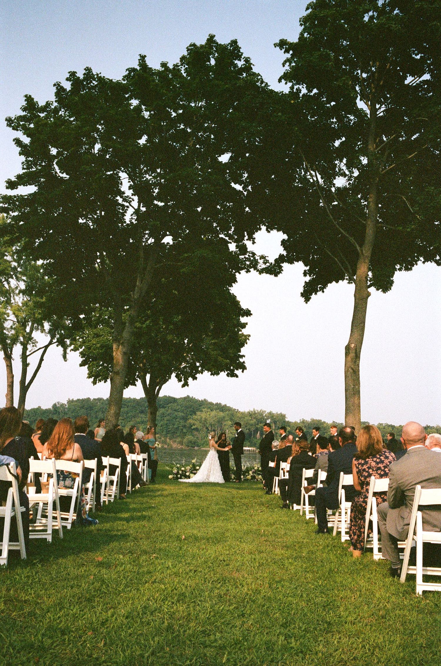 An outdoor wedding ceremony setup with white chairs arranged in rows under tall green trees on a scenic hillside.