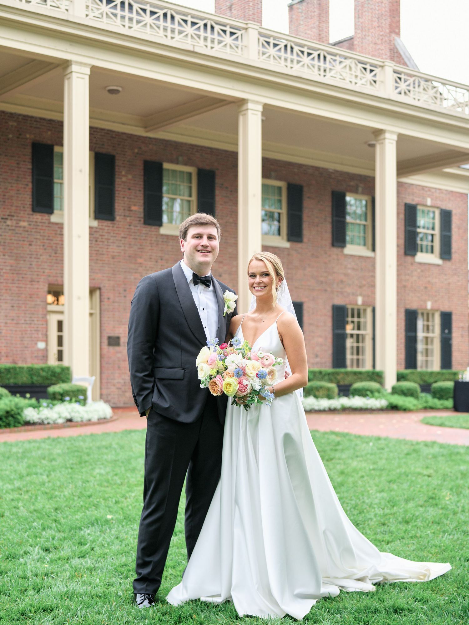 Wedding couple poses in front of a grand brick mansion with white columns and manicured gardens.