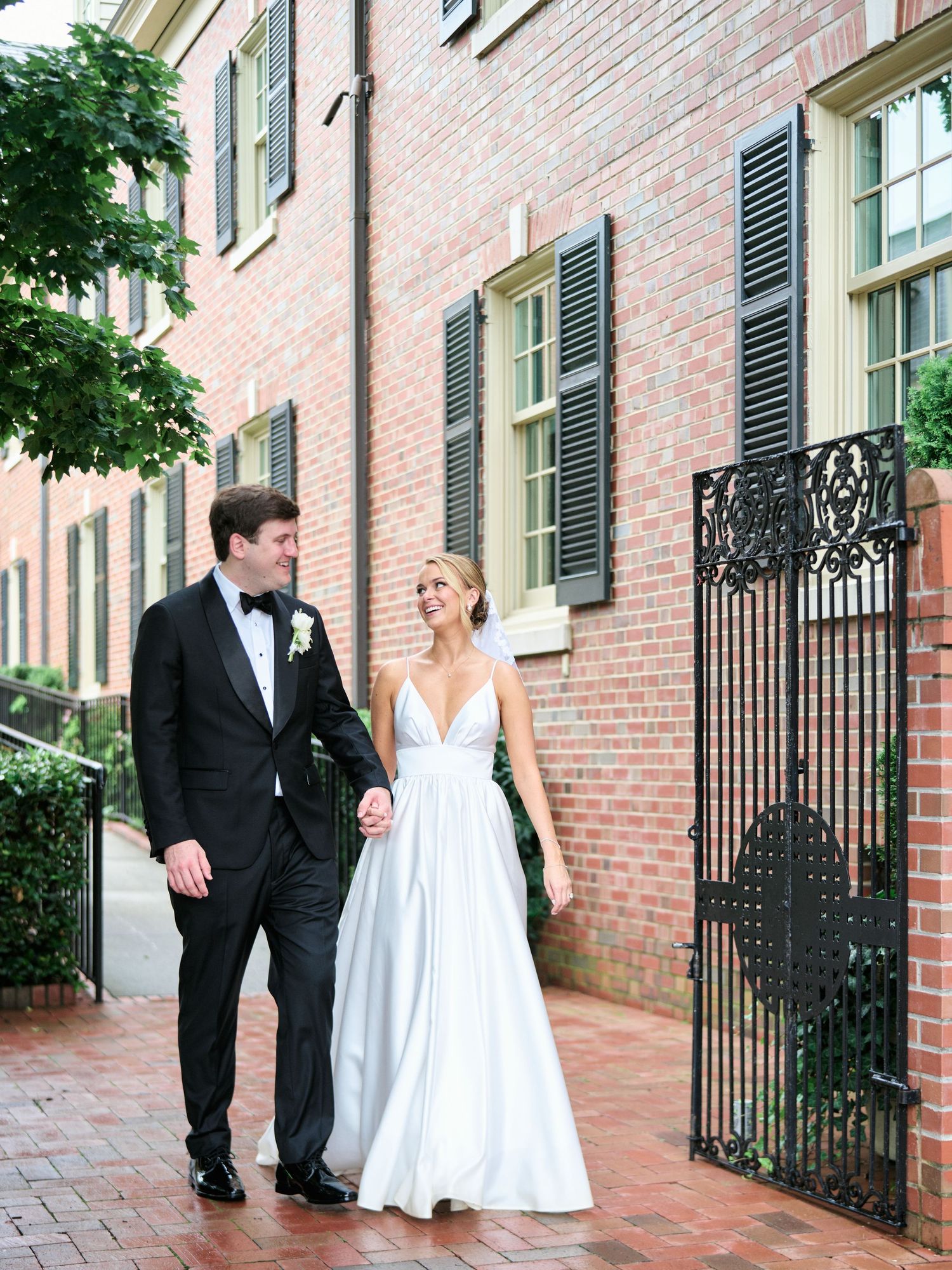 Newlyweds walking along a brick pathway beside an elegant building with black shutters.