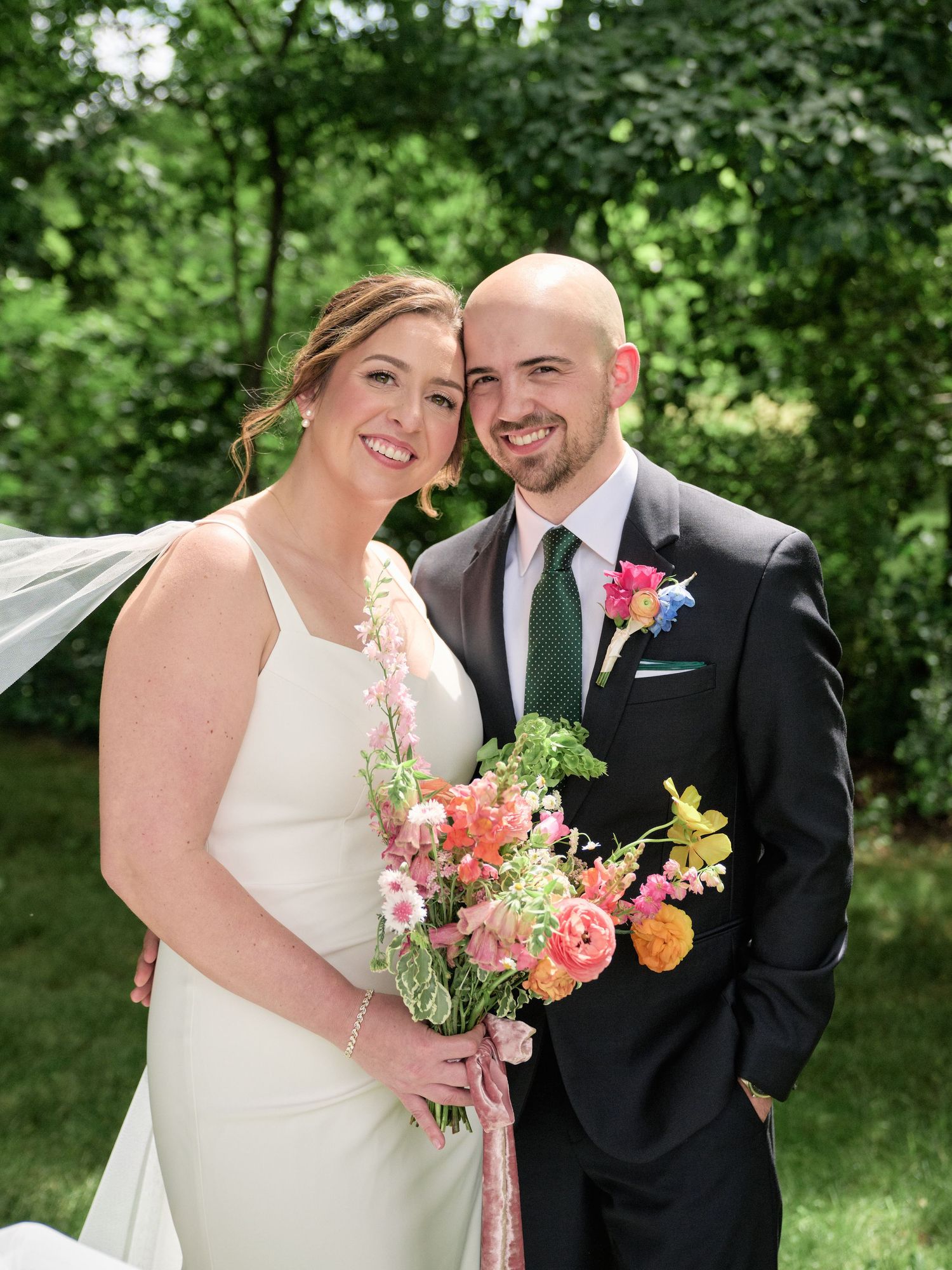 Happy couple shares genuine smiles while posing with coral and yellow bouquet in outdoor setting.