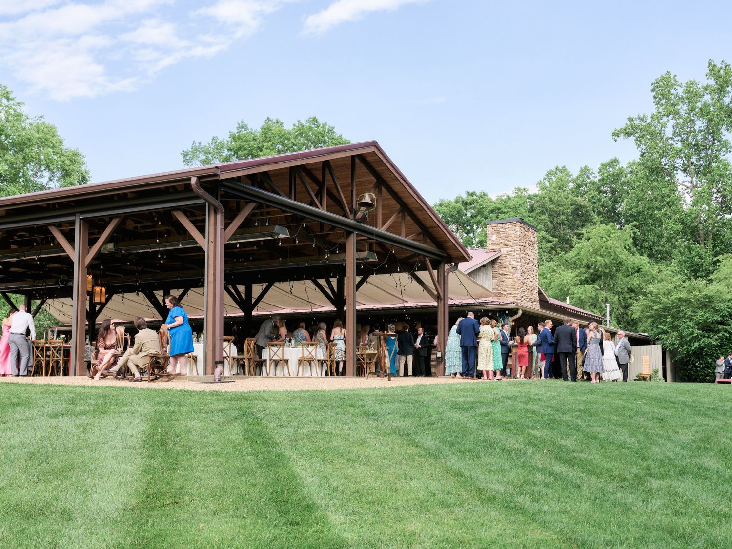 A rustic wooden pavilion with guests gathered on a manicured lawn during an outdoor event.