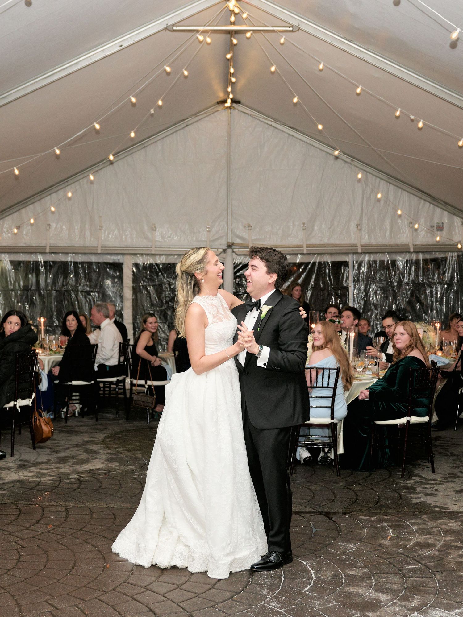 Bride and groom share first dance under a white tent with string lights at elegant wedding reception.