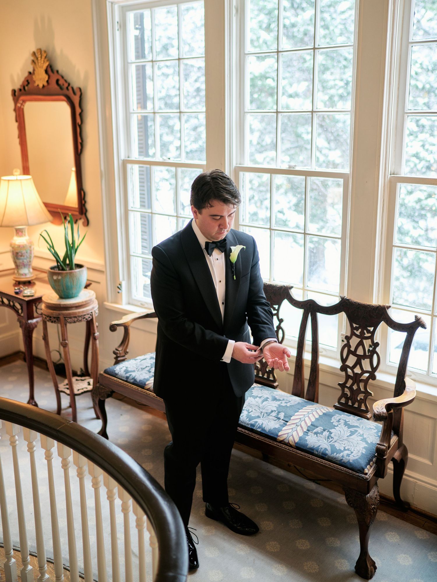 Groom in black tuxedo adjusting cufflinks near elegant windows with antique bench and mirror in a classic interior.