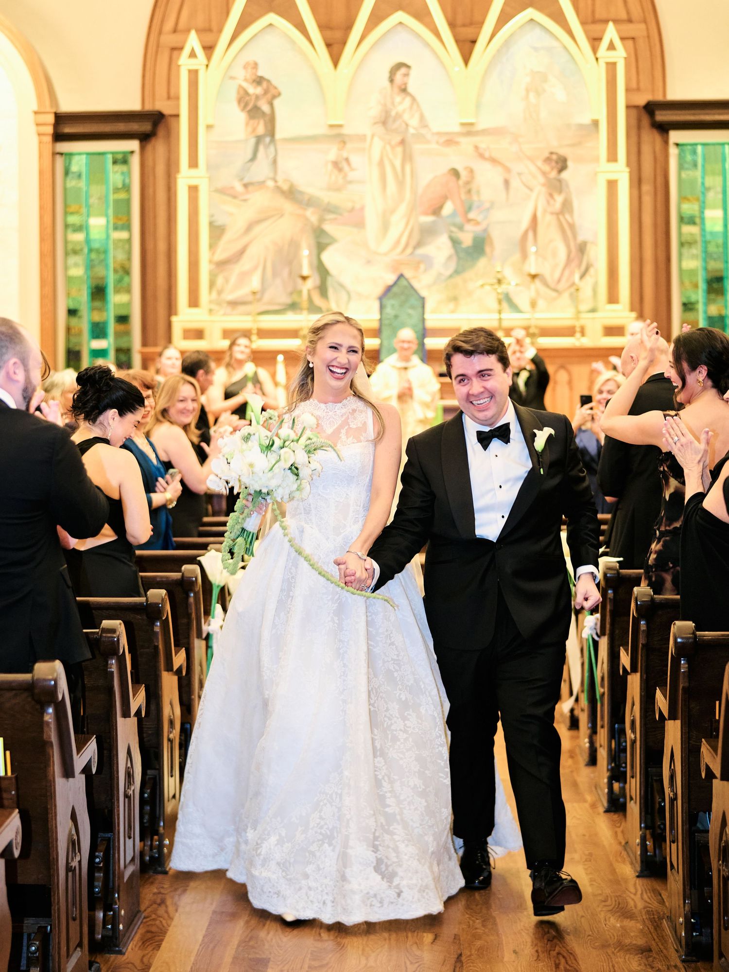 Bride and groom walk down church aisle after wedding ceremony, bride holding white floral bouquet in elegant lace gown.
