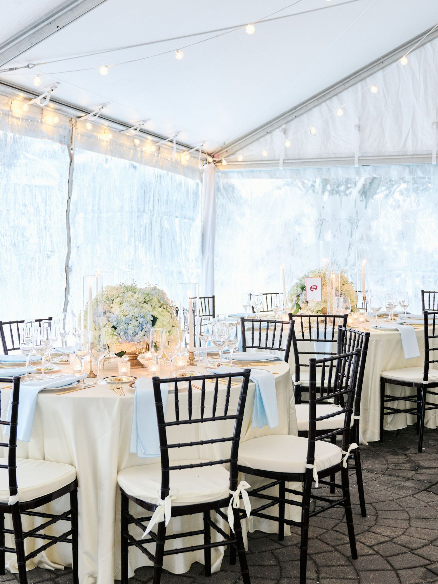 Elegant wedding reception under a white tent with black chiavari chairs, ivory linens, and blue hydrangea centerpieces with string lights.