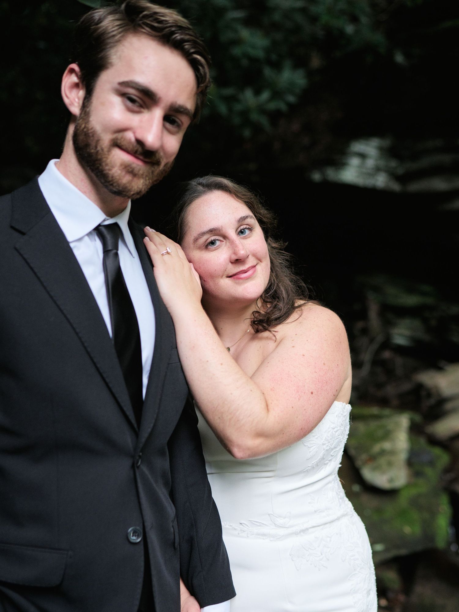 A bride and groom pose together outdoors, she in a white strapless gown and he in a black suit with tie.