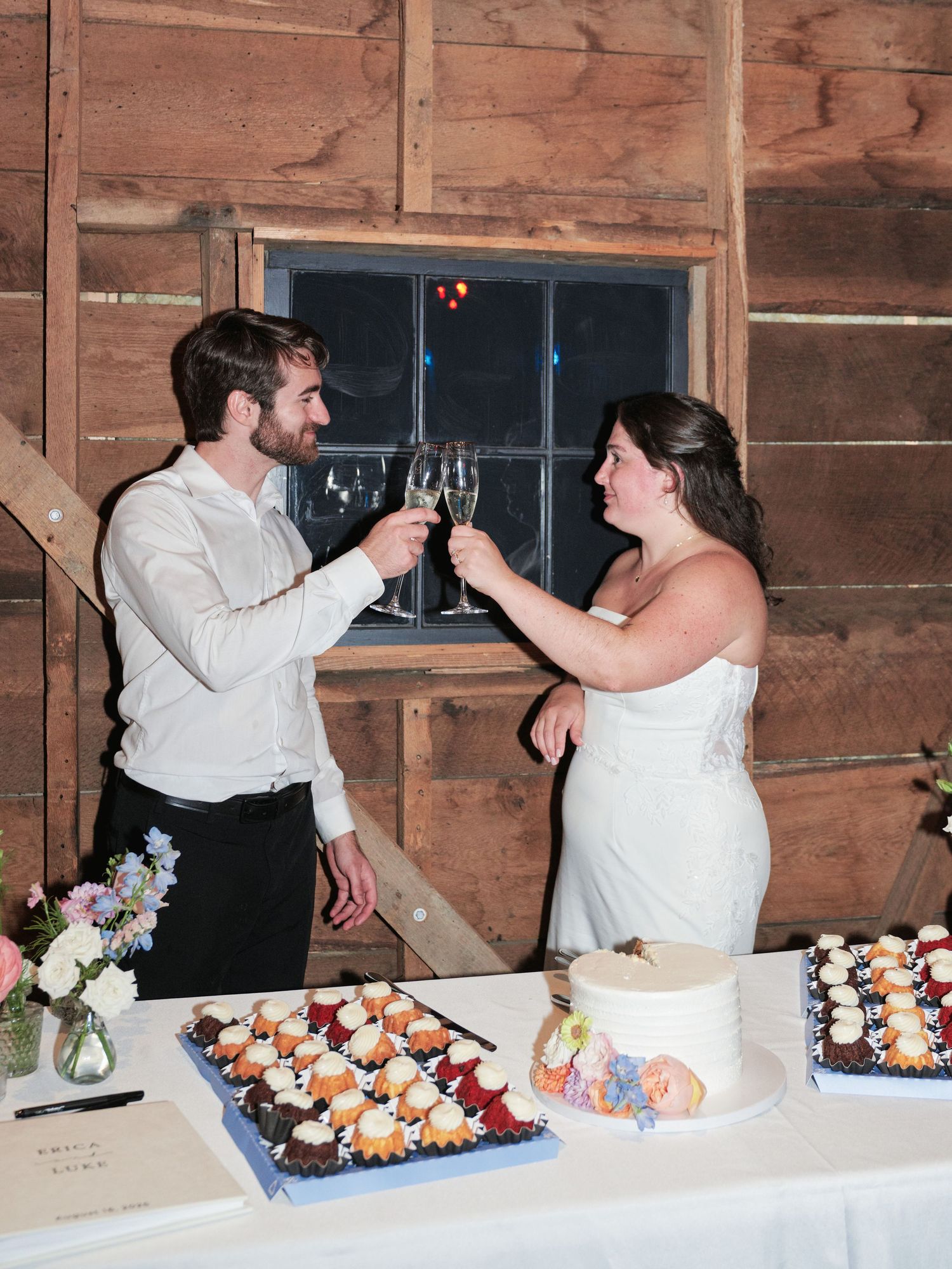 Bride and groom toasting with champagne glasses at rustic barn wedding reception with cupcake dessert table.