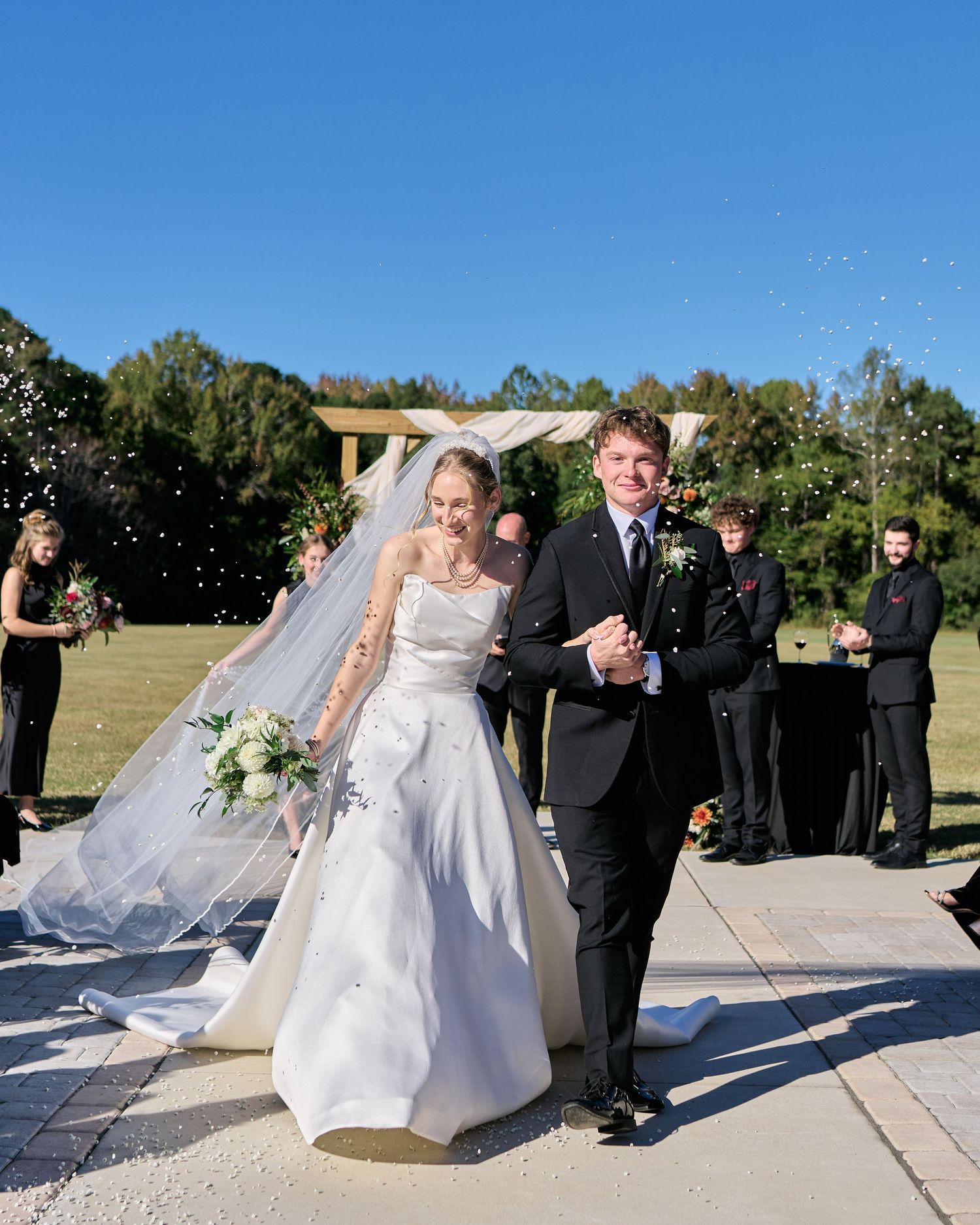A couple celebrates their wedding ceremony outdoors on a sunny day with guests and confetti in the background.