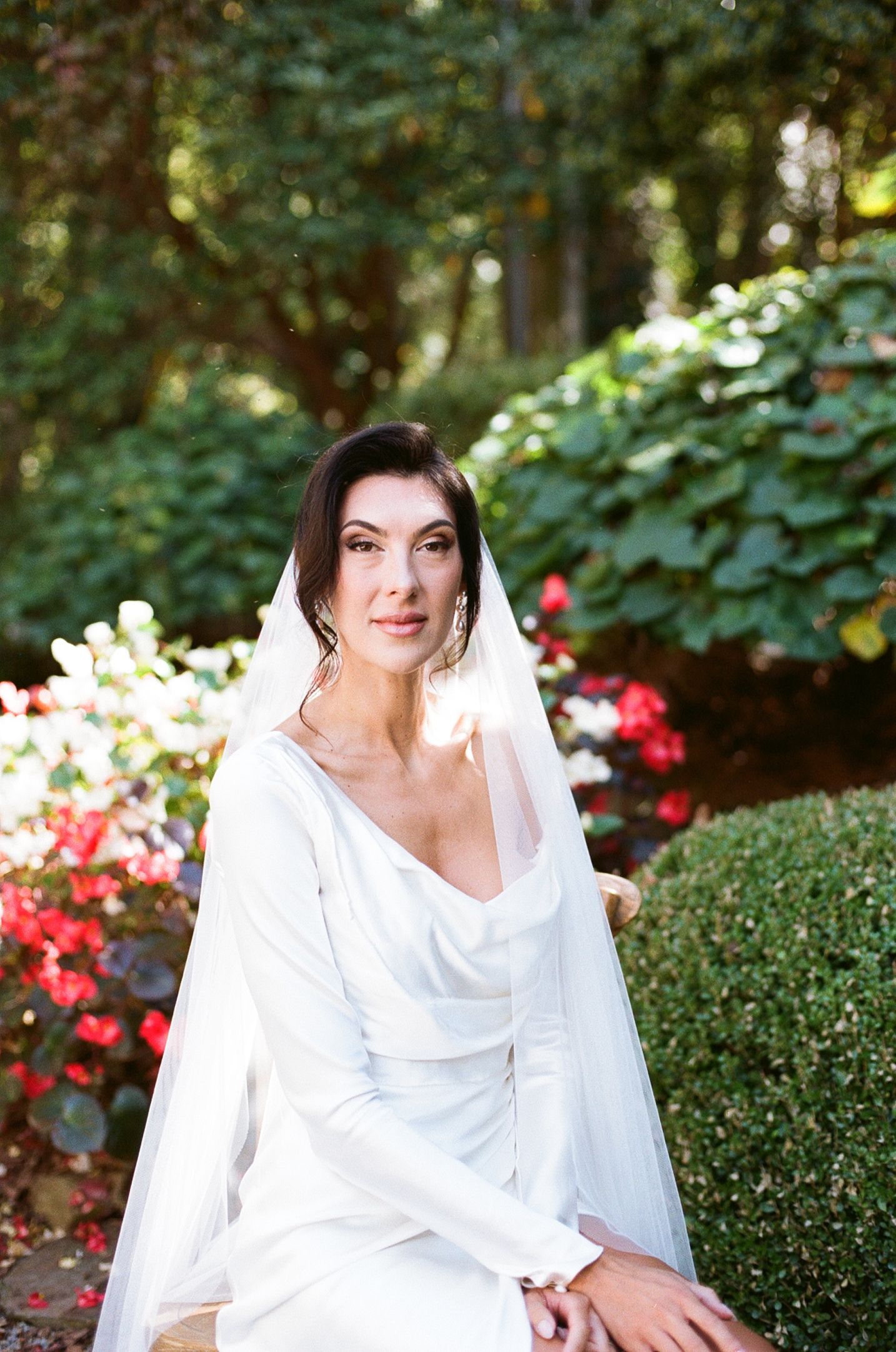 A bride in a classic white long-sleeved wedding gown and veil poses in a garden with red flowers and greenery.