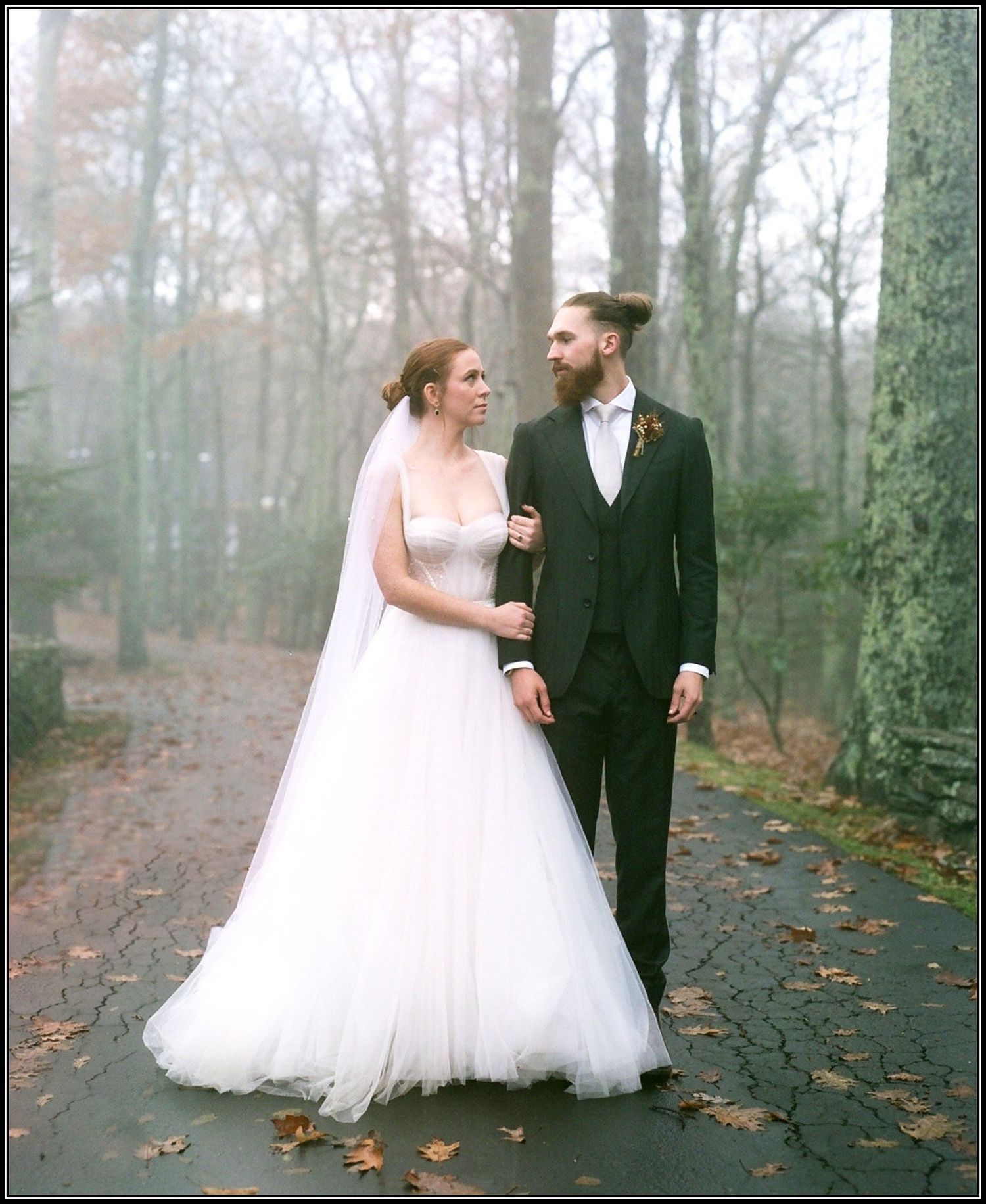 A couple in wedding attire walks along a misty autumn path covered in fallen leaves.