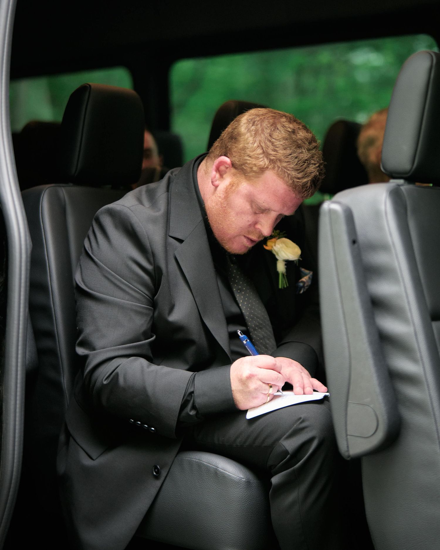 A person in a suit sits in a car writing on paper while holding a pen.