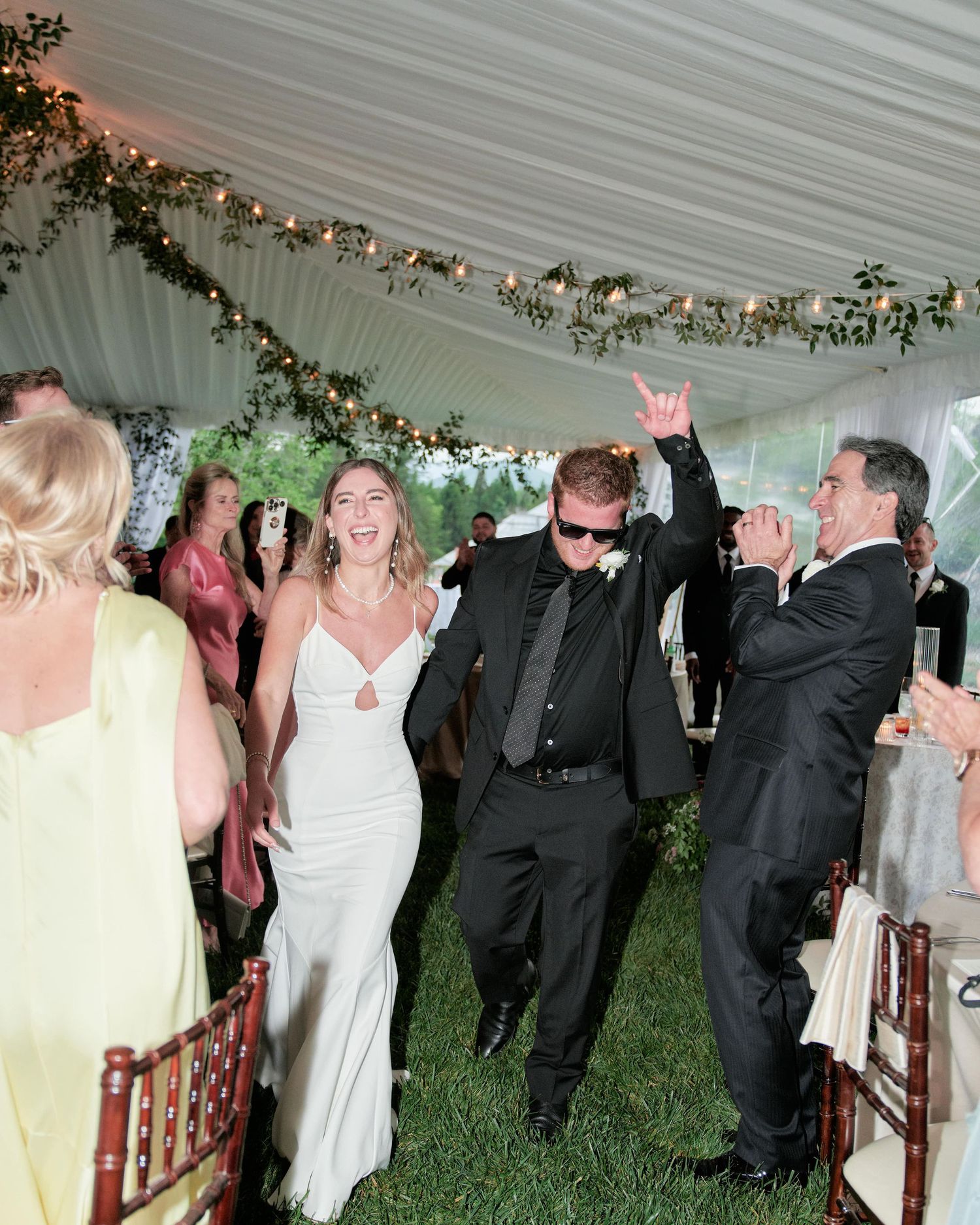 Wedding guests dance and celebrate together under a white tent decorated with floral garlands during an evening reception.