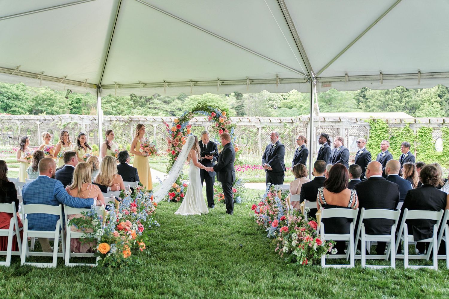 Wedding guests seated under white tent canopy during outdoor ceremony react with joyful expressions and gestures.