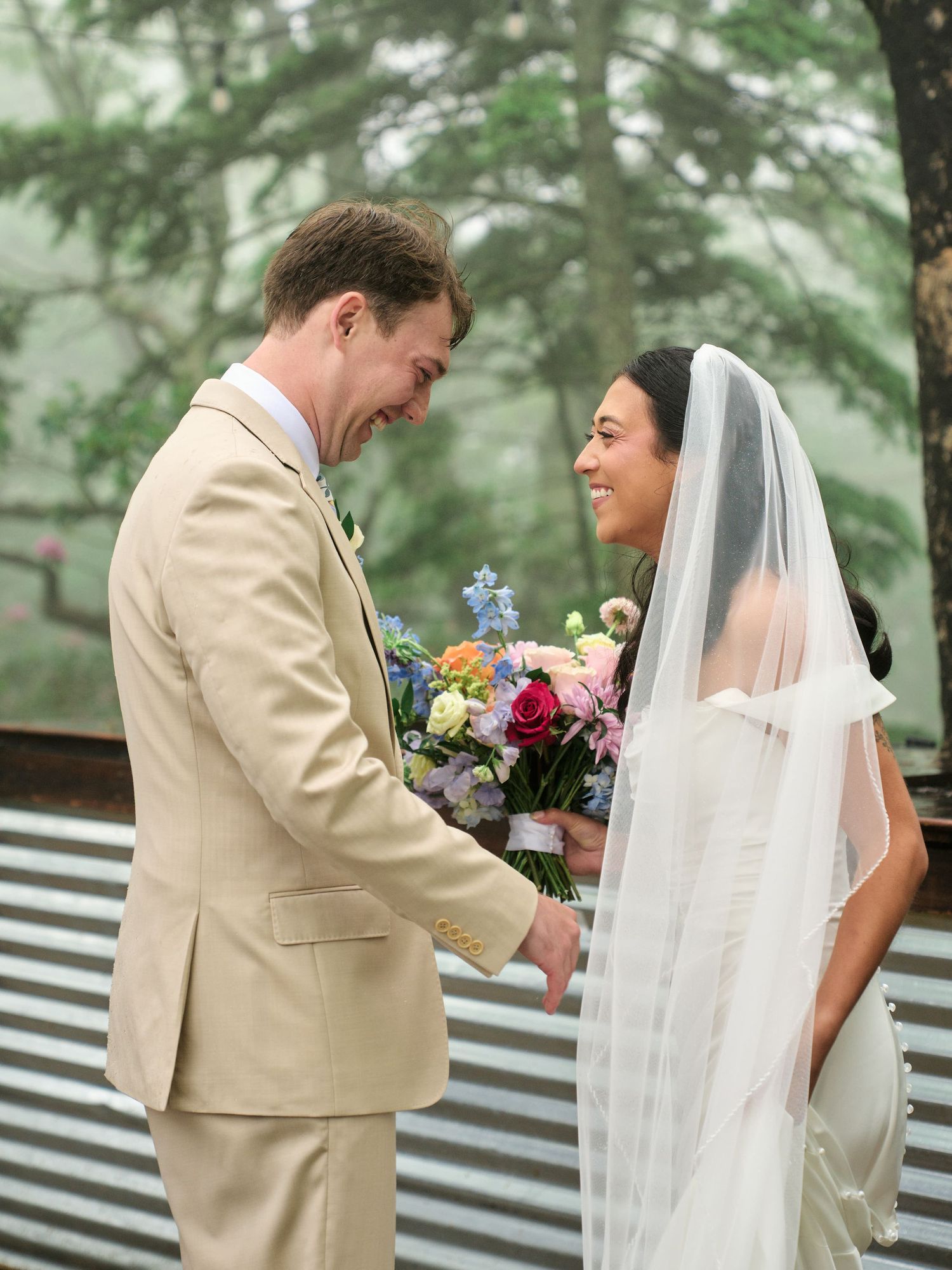 Smiling bride in white gown and veil holds colorful bouquet with groom in tan suit during outdoor wedding ceremony.