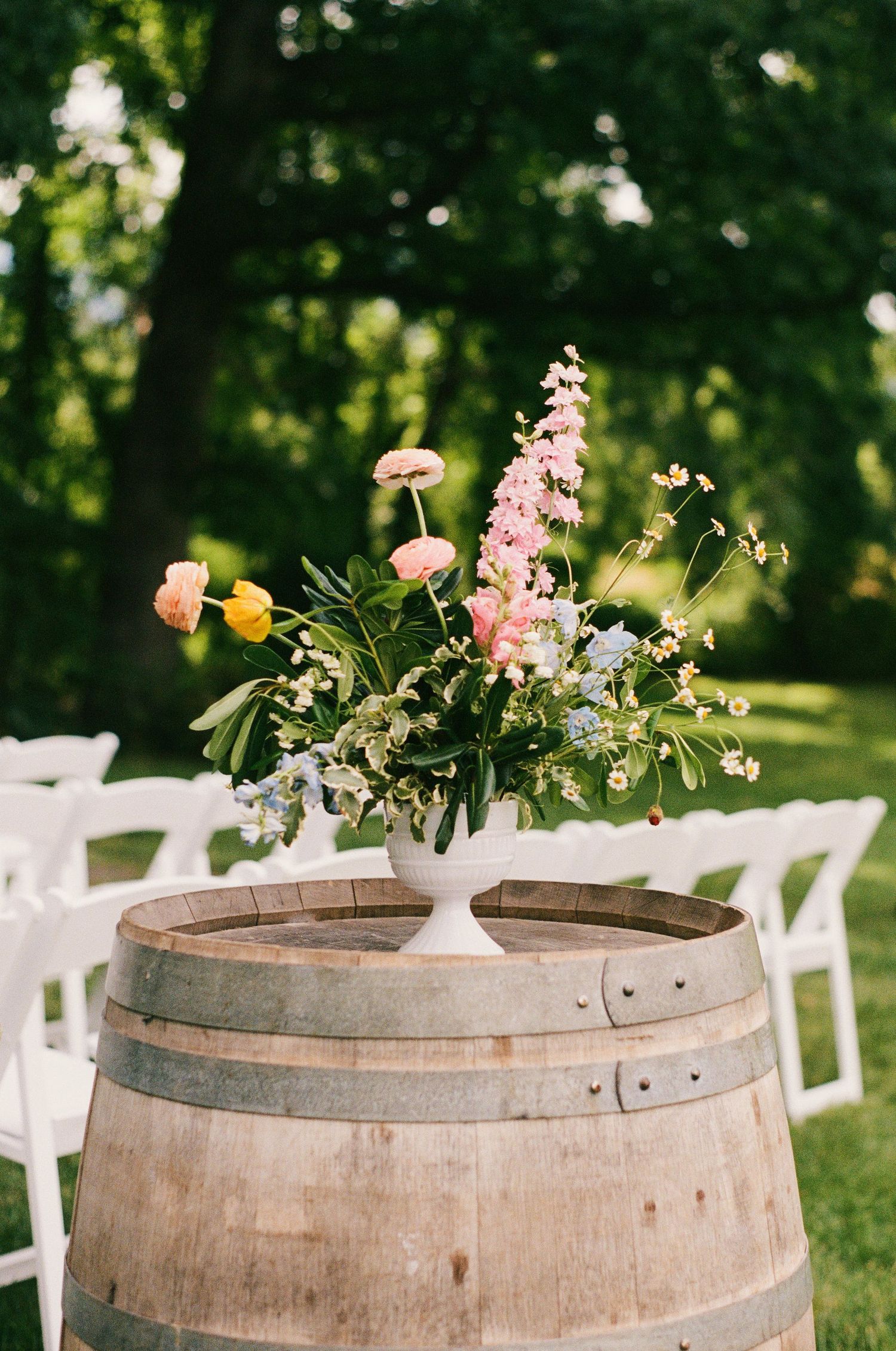 Floral arrangement in white vase on rustic wine barrel at outdoor wedding ceremony with white chairs.