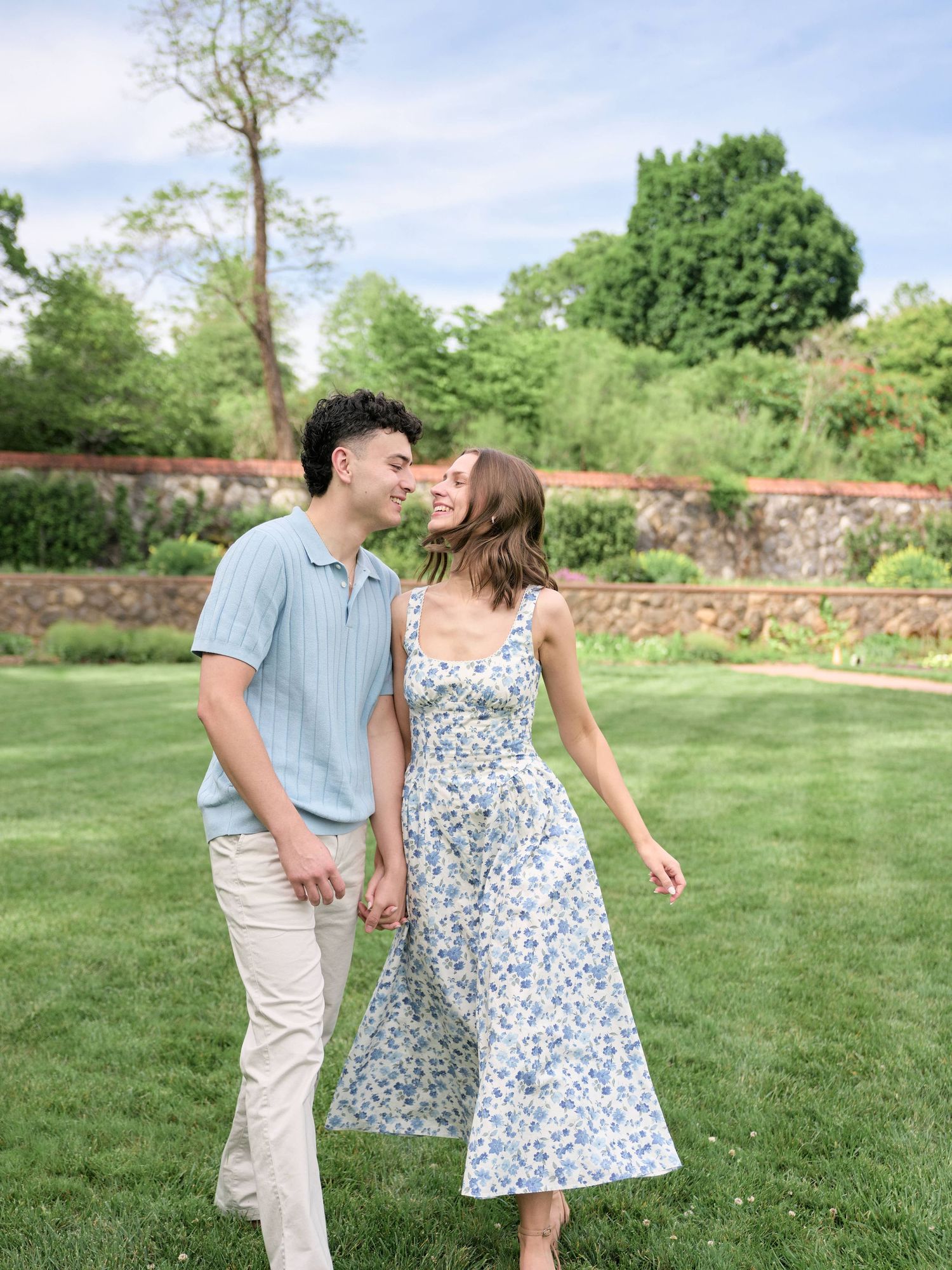 A couple walks hand-in-hand on lush green grass, sharing a kiss, with a stone wall and trees in the background.