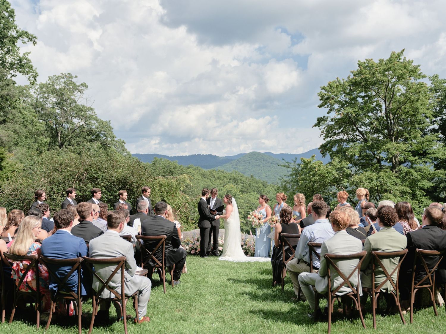 Outdoor wedding ceremony on a green lawn with mountain views, guests seated in wooden chairs facing the couple.