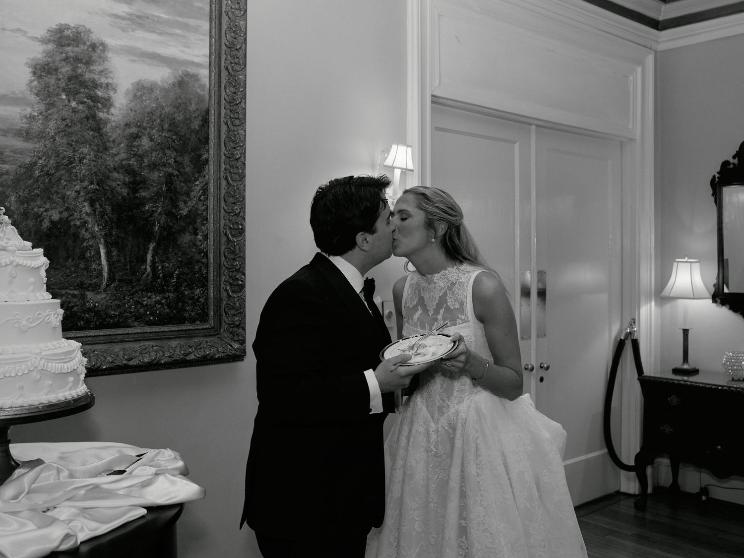 Black and white photo of a bride and groom sharing a kiss near a wedding cake in an elegant room.