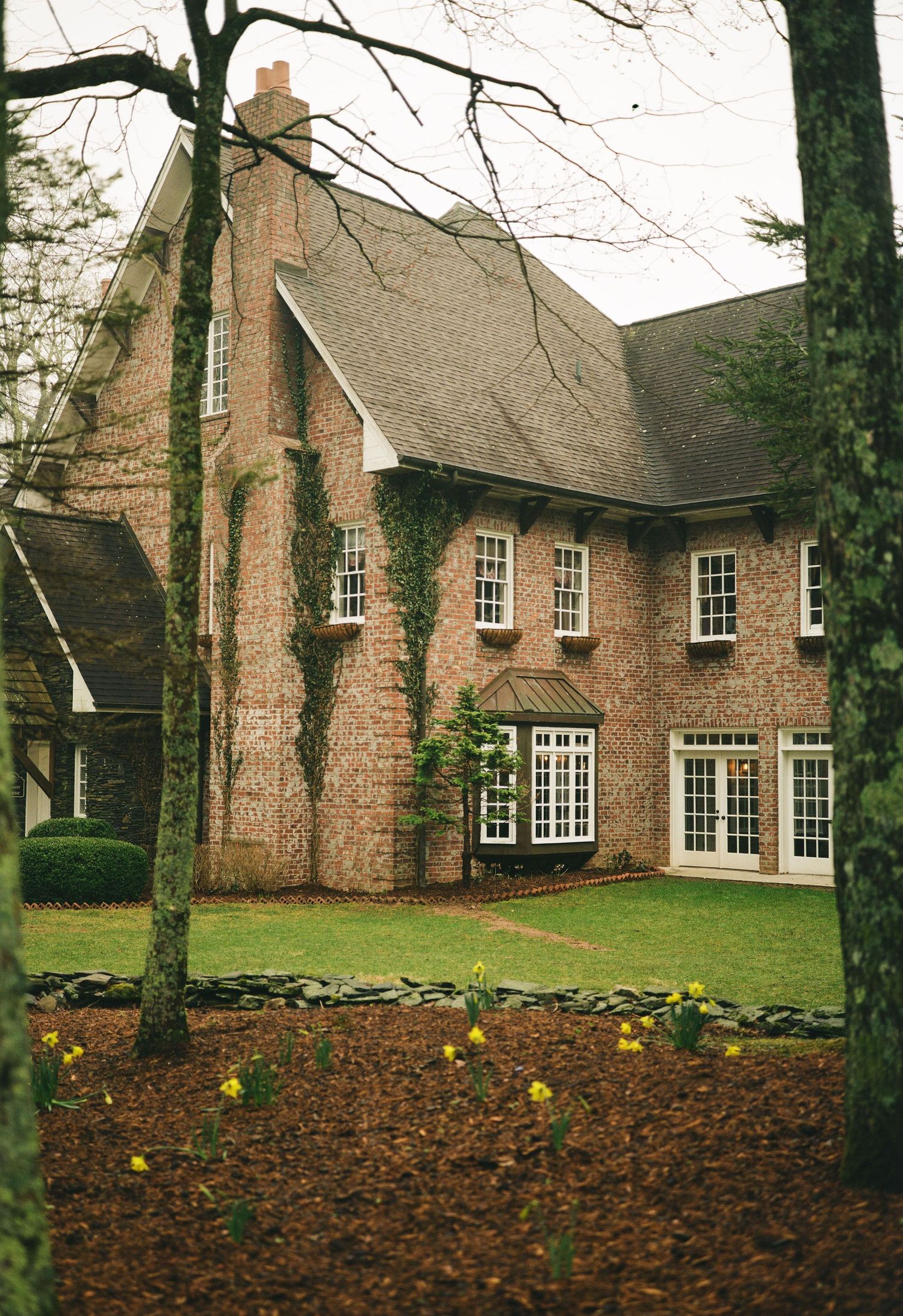 Brick two-story home with ivy-covered walls, white French doors, and daffodils blooming near tree-lined lawn.