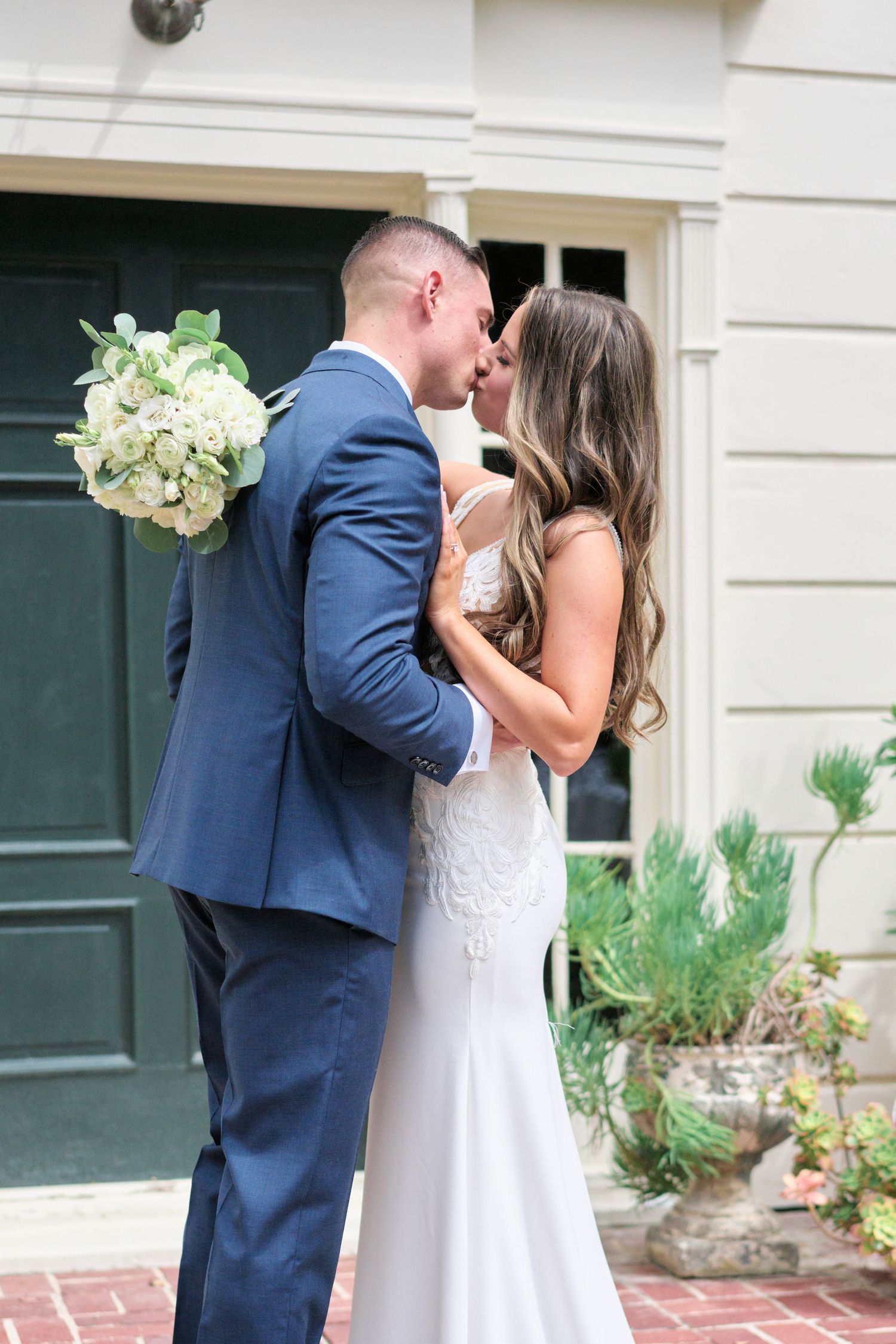 Newlyweds sharing a romantic kiss in front of a dark green door while holding a white floral bouquet.