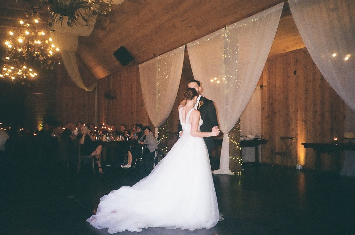 Bride and groom dancing under chandelier in rustic venue with dramatic lighting and white drapes.