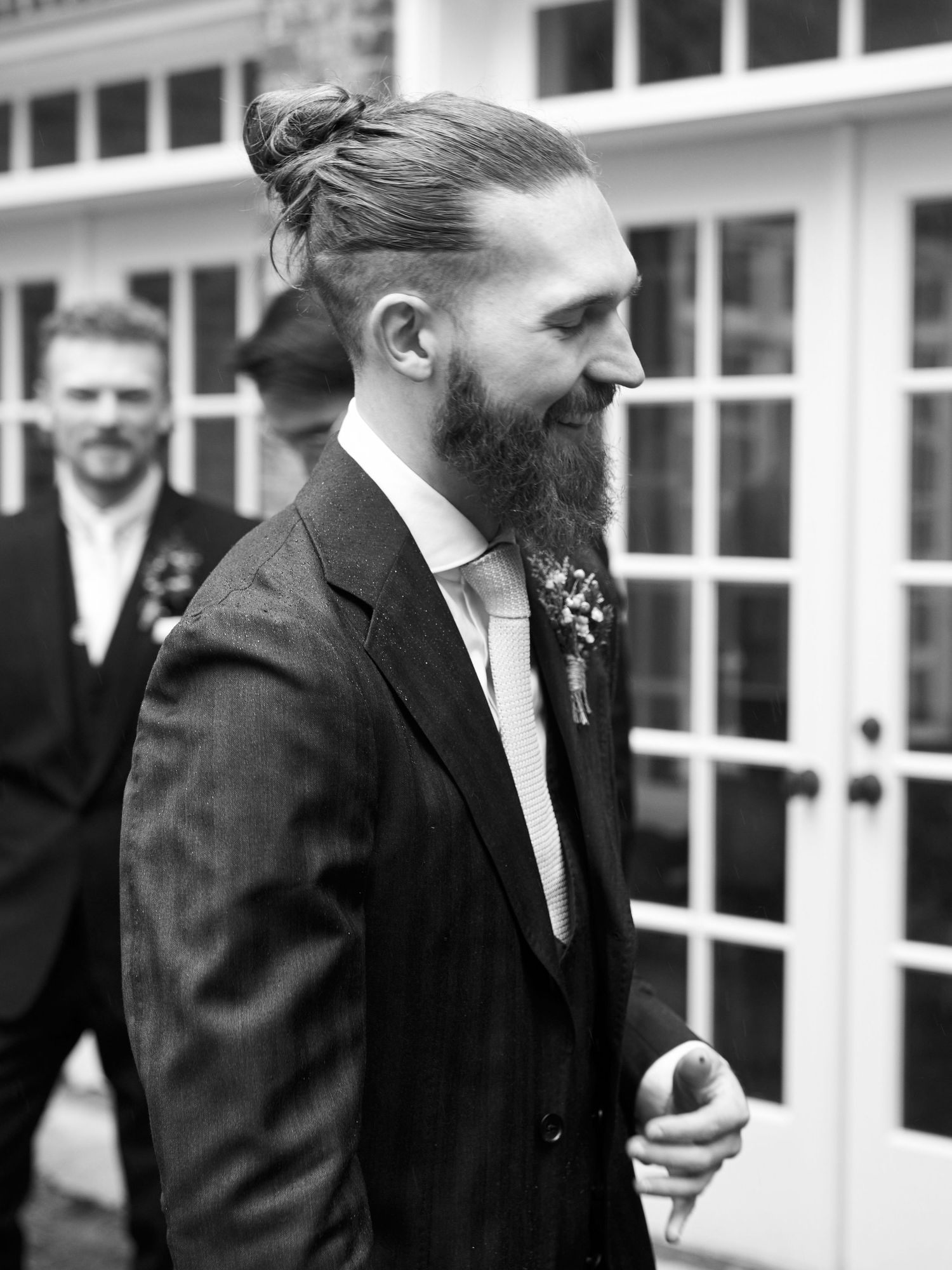 Stylish suited person with beard standing outdoors near French doors.