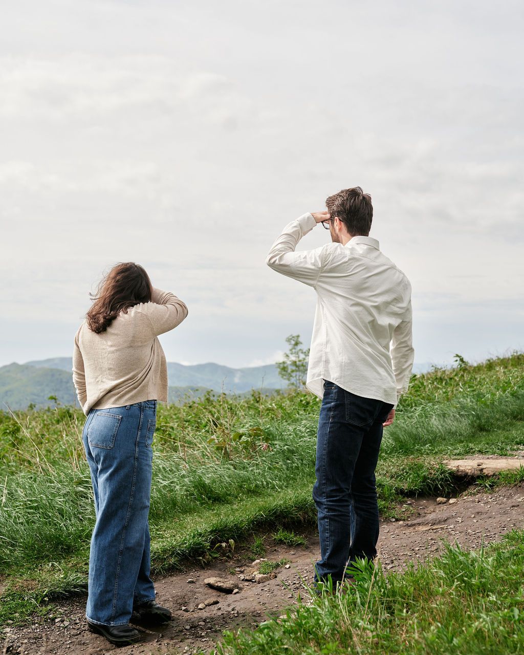 Two people stand on a grassy hilltop looking into the distance at a mountain landscape.