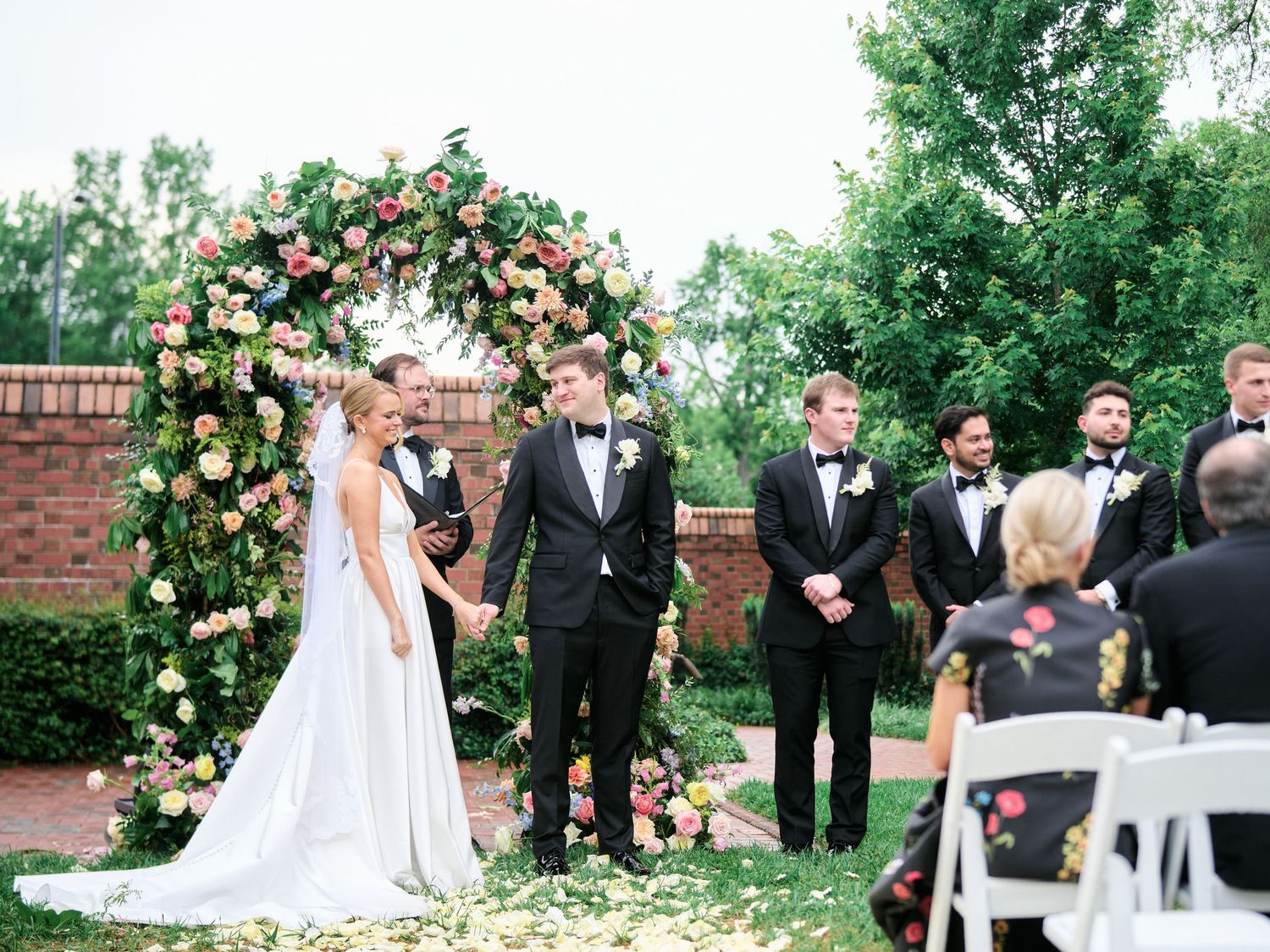 Outdoor wedding ceremony under a beautiful floral arch with pink and cream roses against a brick wall backdrop.