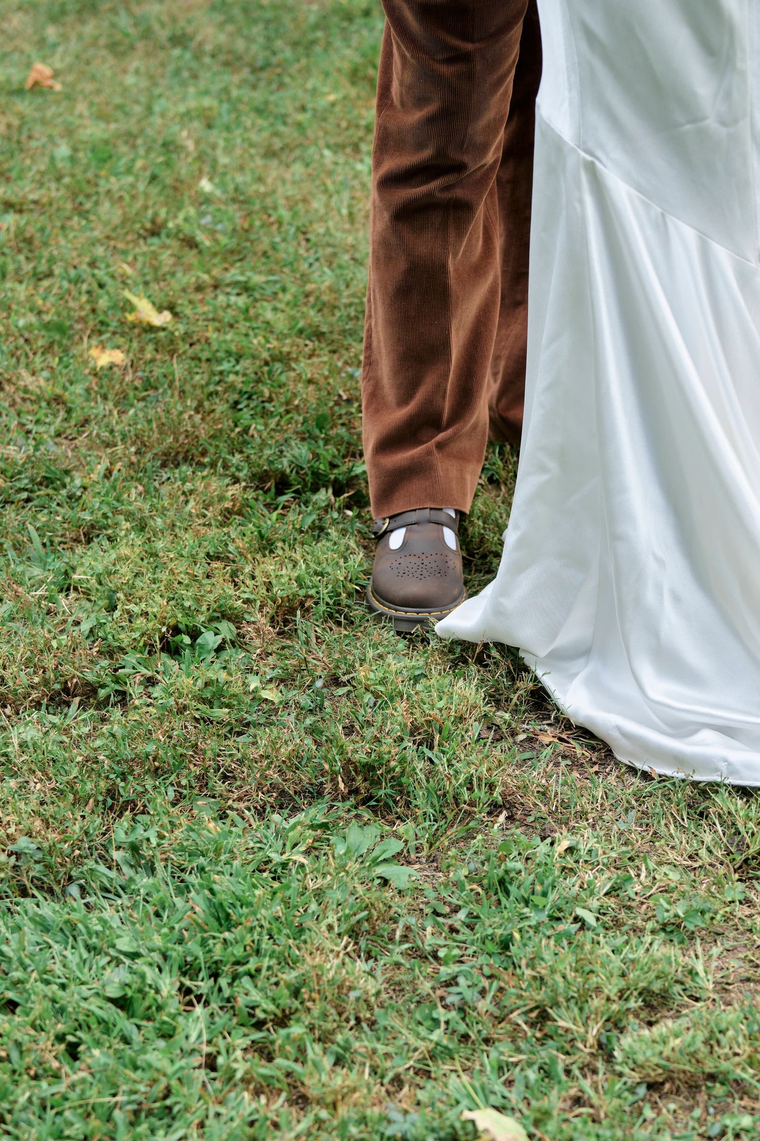 Detail shot of brown dress shoes and white wedding dress hem on grass.
