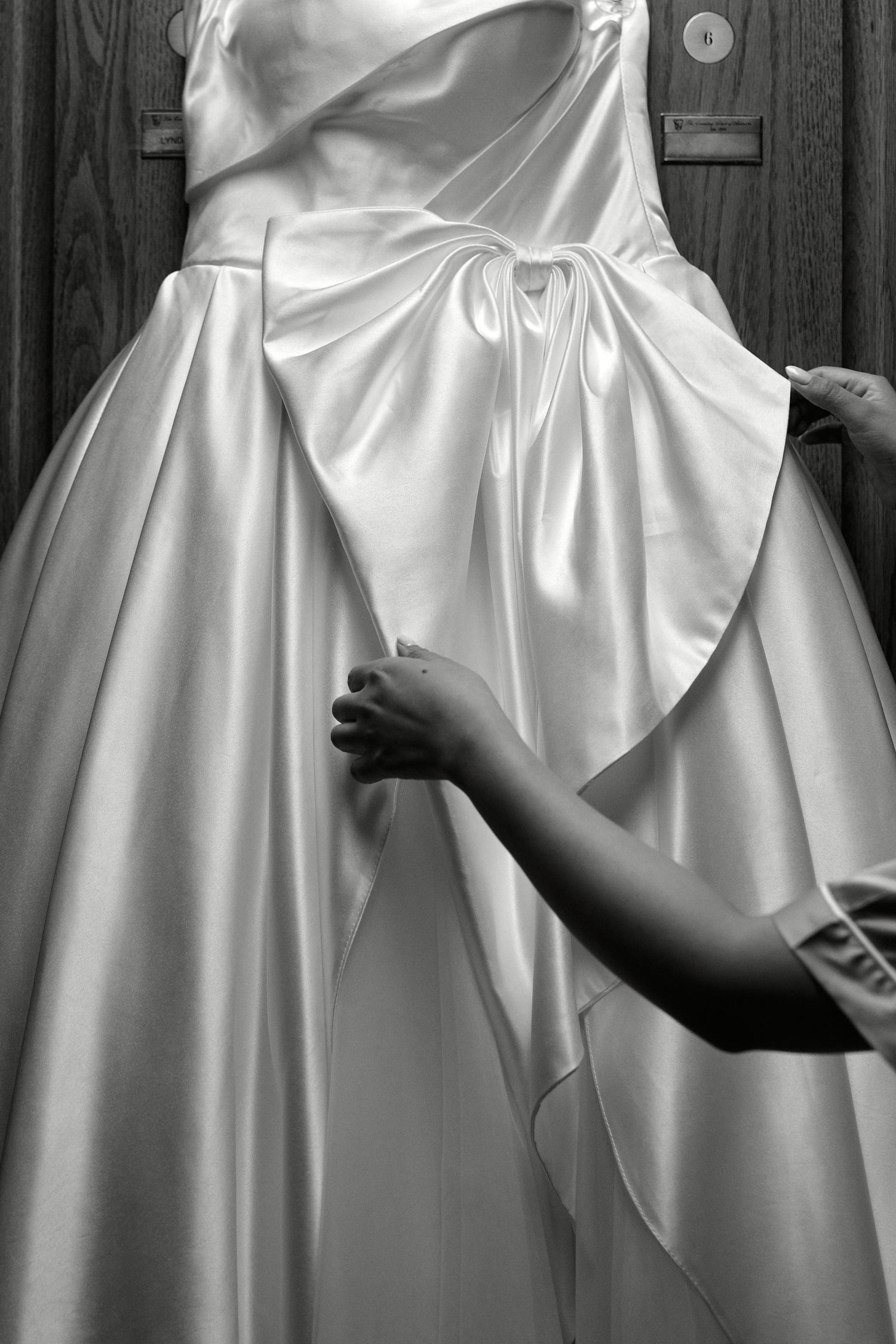 Close-up black and white detail of hands adjusting the folds of a satin wedding dress.