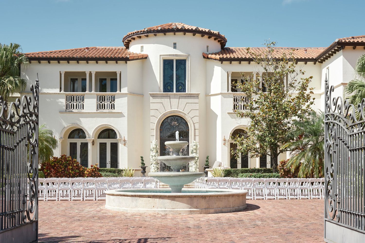 Mediterranean-style wedding venue with white facade, arched fountain and ceremony chairs arranged in courtyard.