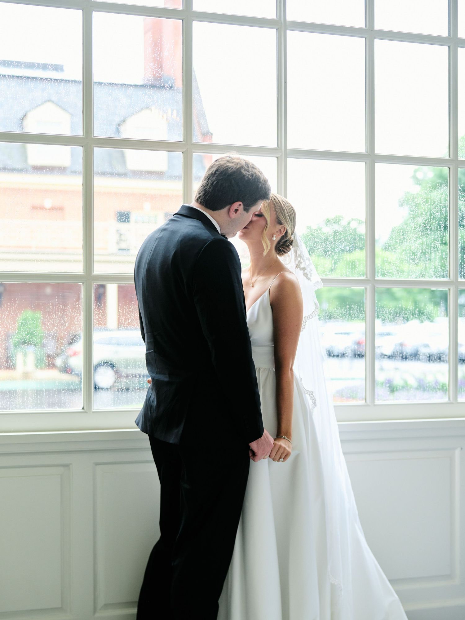 Couple embracing by large window in classic wedding attire.