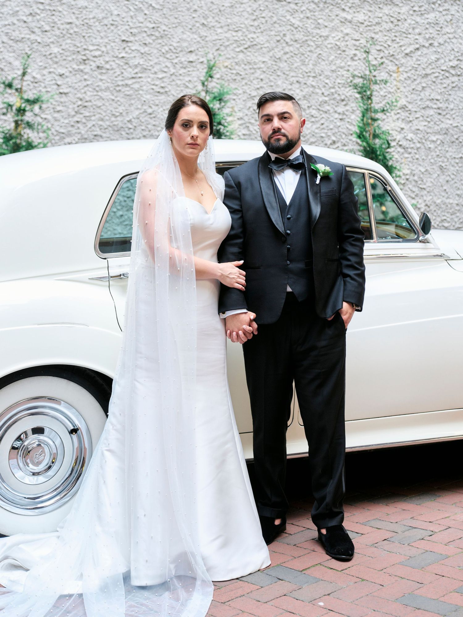 A bride and groom pose formally beside a vintage white car on their wedding day.