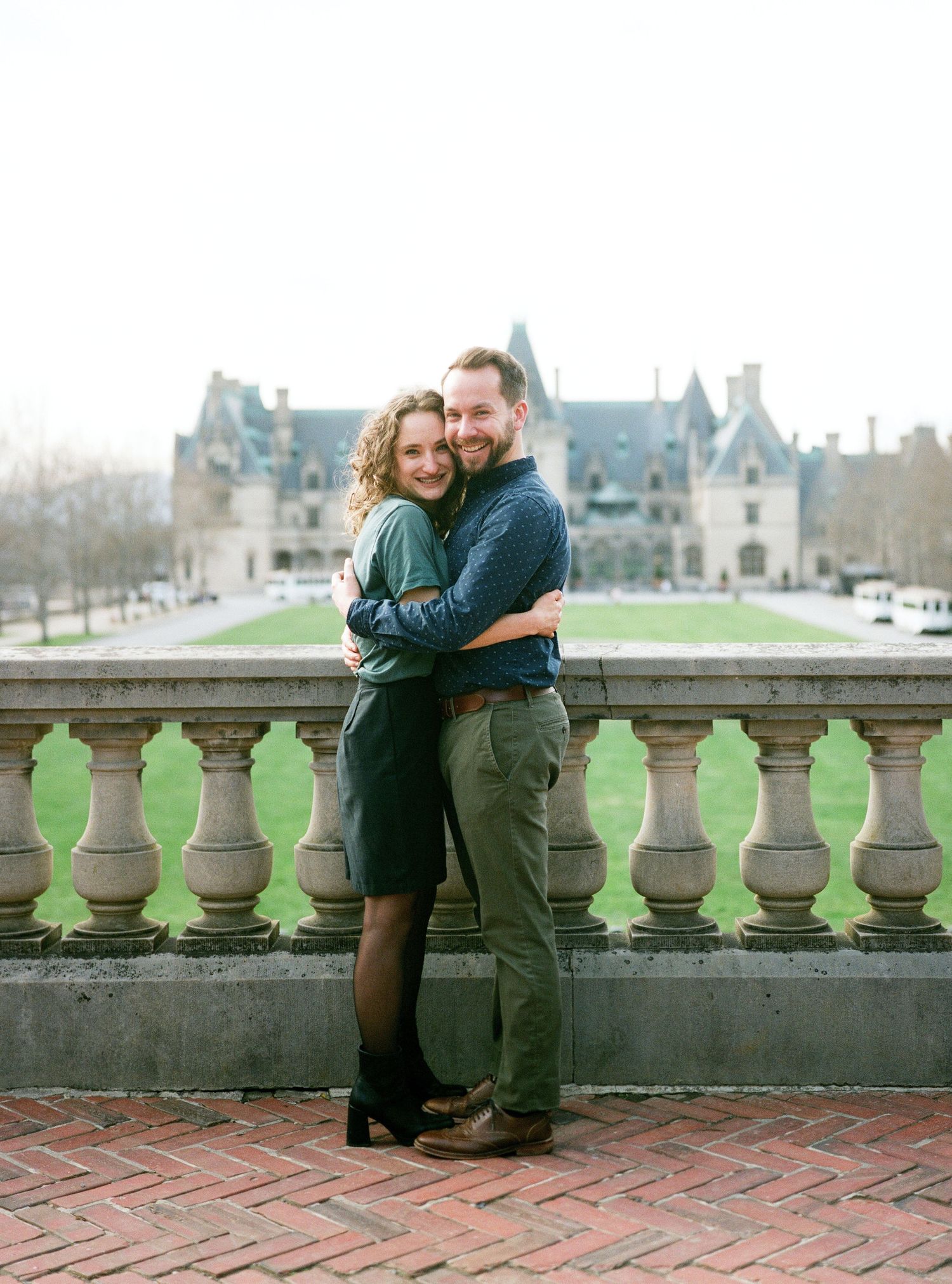 Happy couple embracing on stone balcony with historic mansion in background.