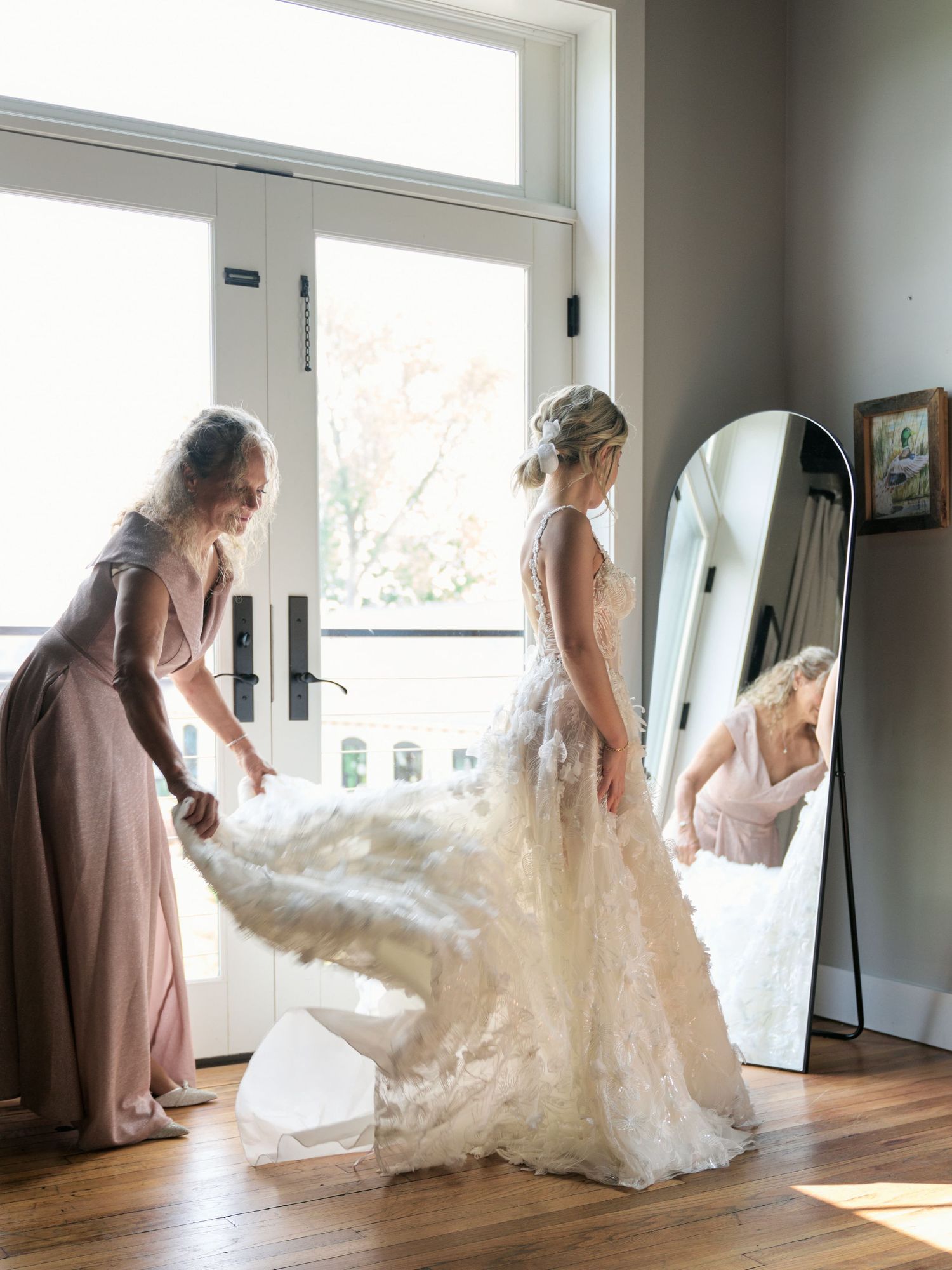A bride in a flowing lace wedding gown gets ready in front of a full-length mirror while helper adjusts the dress.