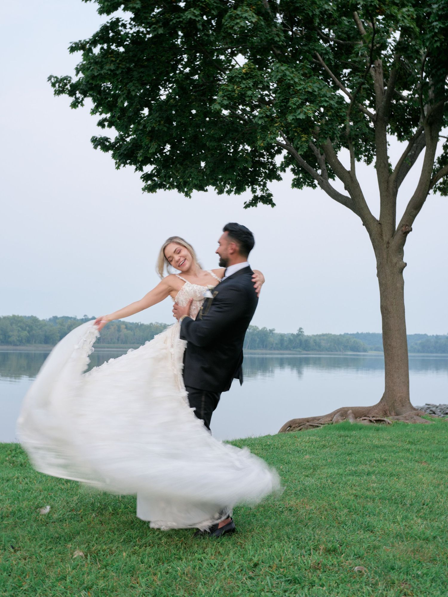 A couple in wedding attire dances joyfully by a lakeside tree, with the bride's dress twirling in a dramatic spin.
