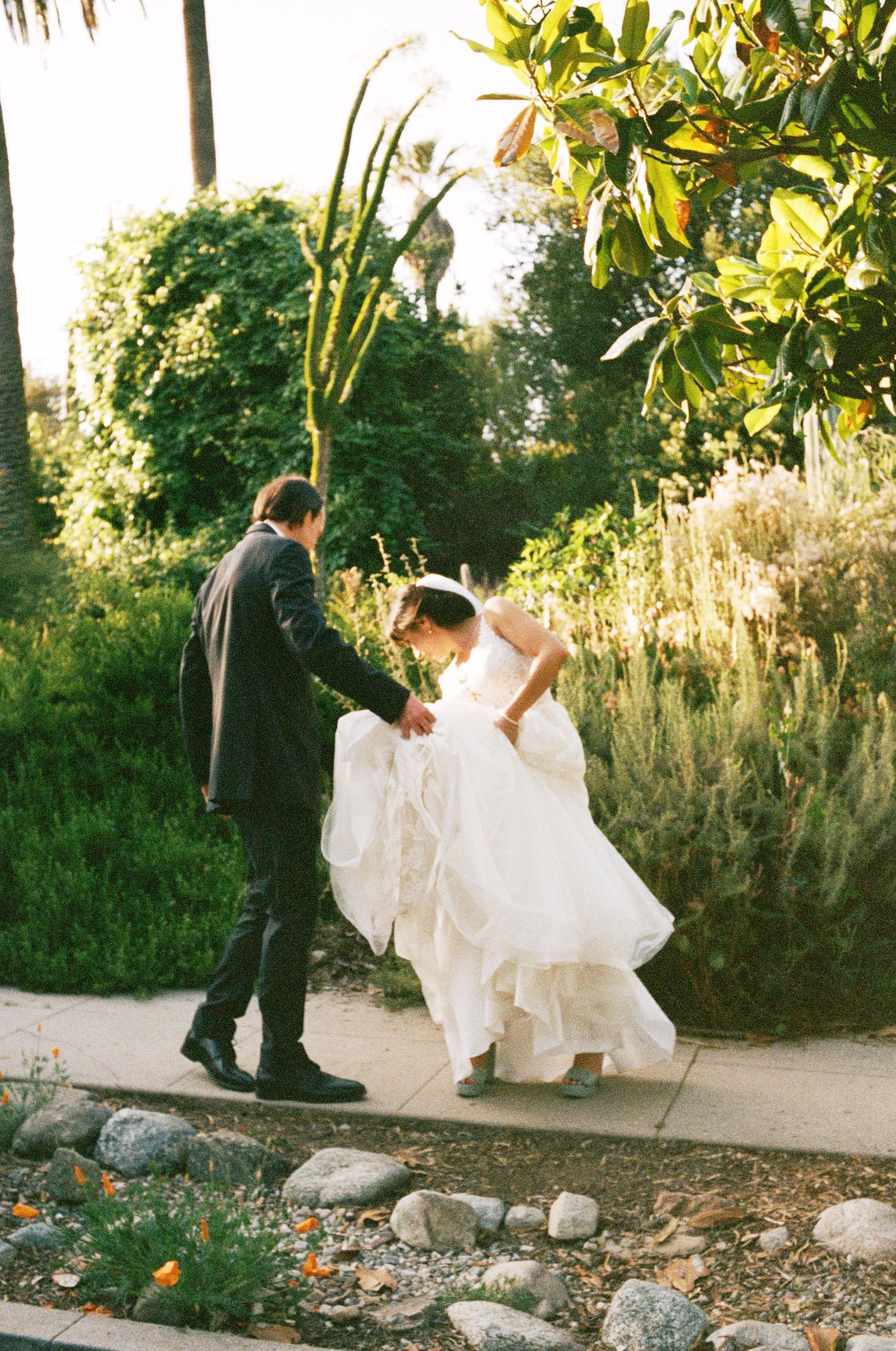 A couple in formal wedding attire walks along a garden path during golden hour.