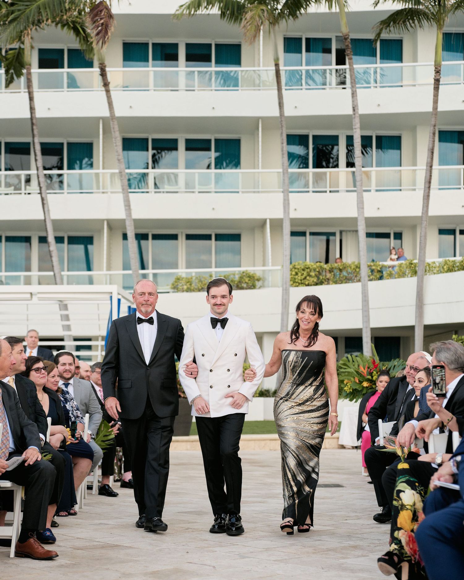 Wedding processional walking down outdoor aisle with palm trees and modern hotel balconies in background.