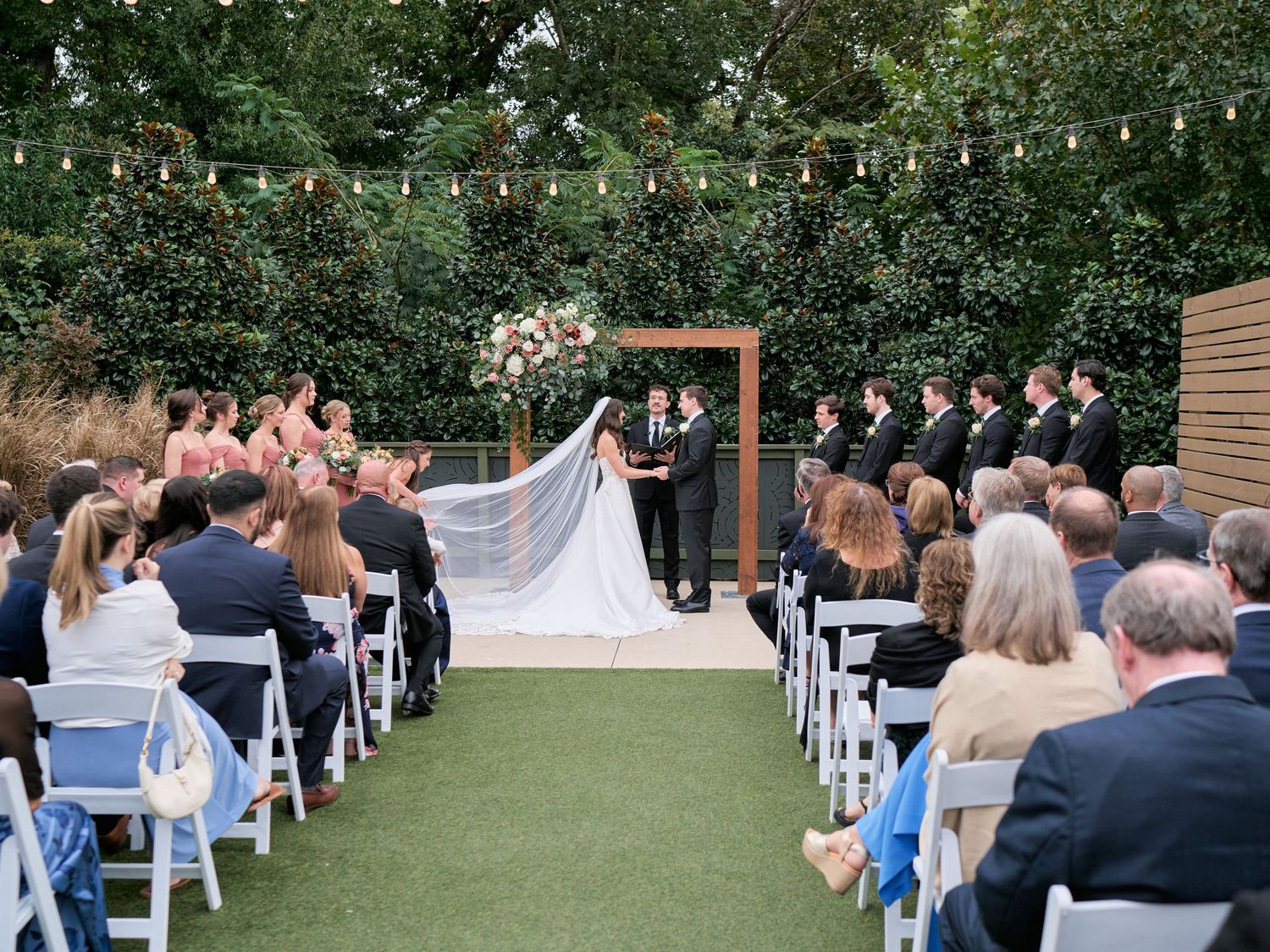Outdoor wedding ceremony setup with white chairs and string lights in a garden setting under trees.