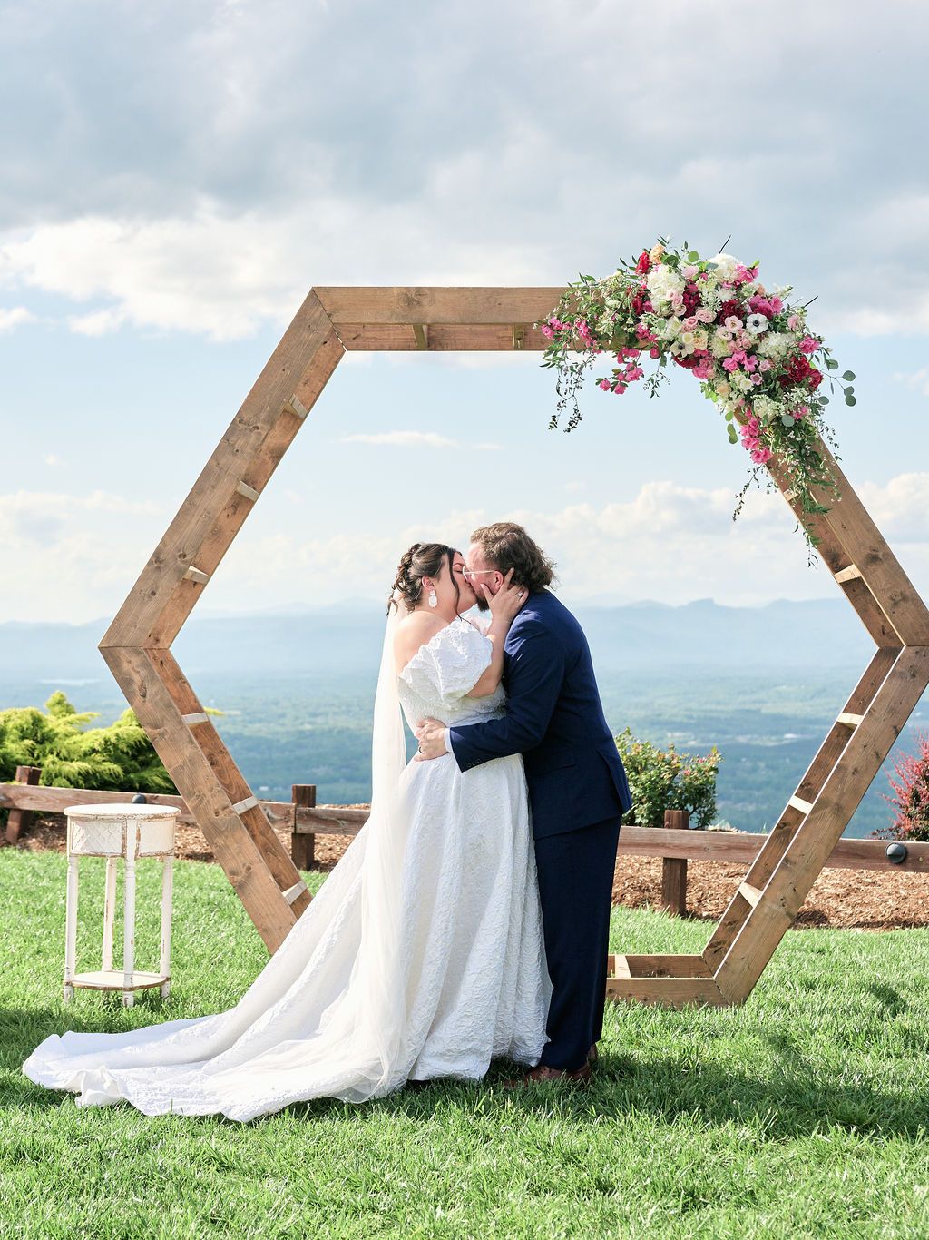 Couple sharing romantic moments on a hilltop with wooden hexagonal arch frames and floral arrangements in scenic setting.