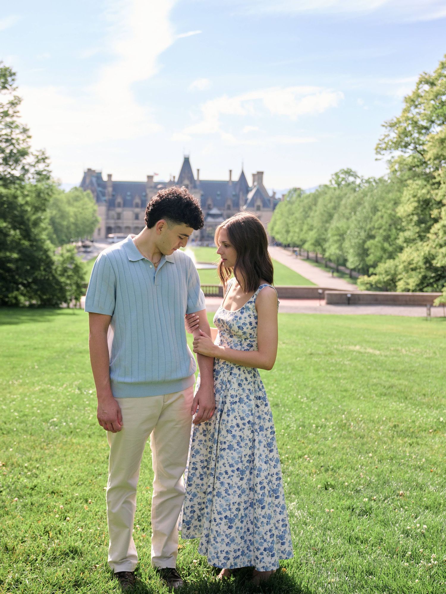 A young couple stands on a green lawn in front of a grand estate on a sunny day, she in a floral dress, he in a blue polo.