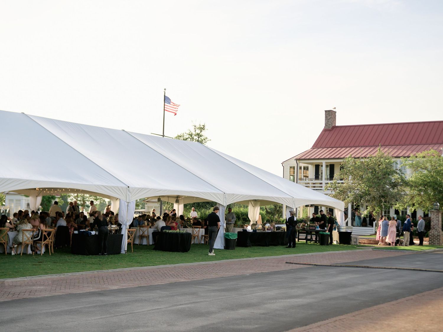 Elegant outdoor wedding reception under large white tent near historic building with American flag at dusk.