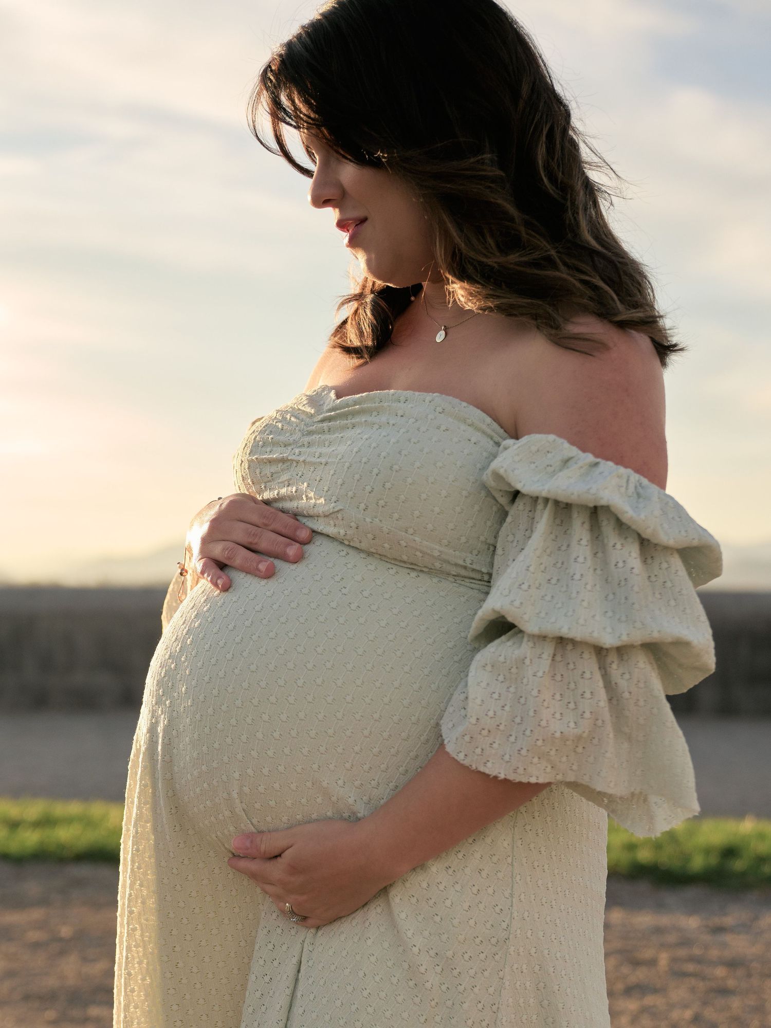 Pregnant woman in off-shoulder cream lace dress cradling baby bump outdoors at golden hour.