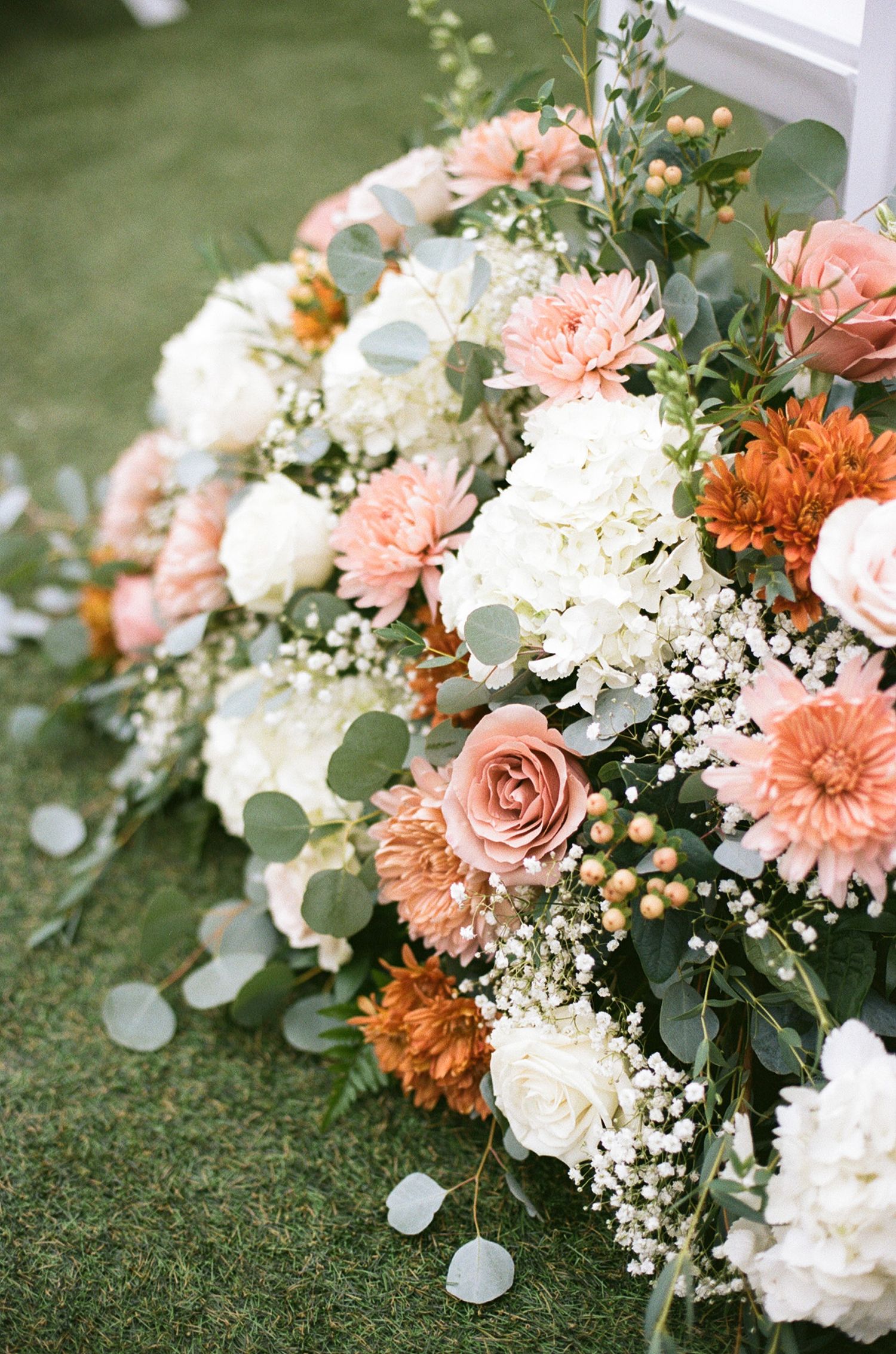 Lush wedding floral arrangement featuring peach roses, white hydrangeas, orange dahlias, and eucalyptus on green grass.