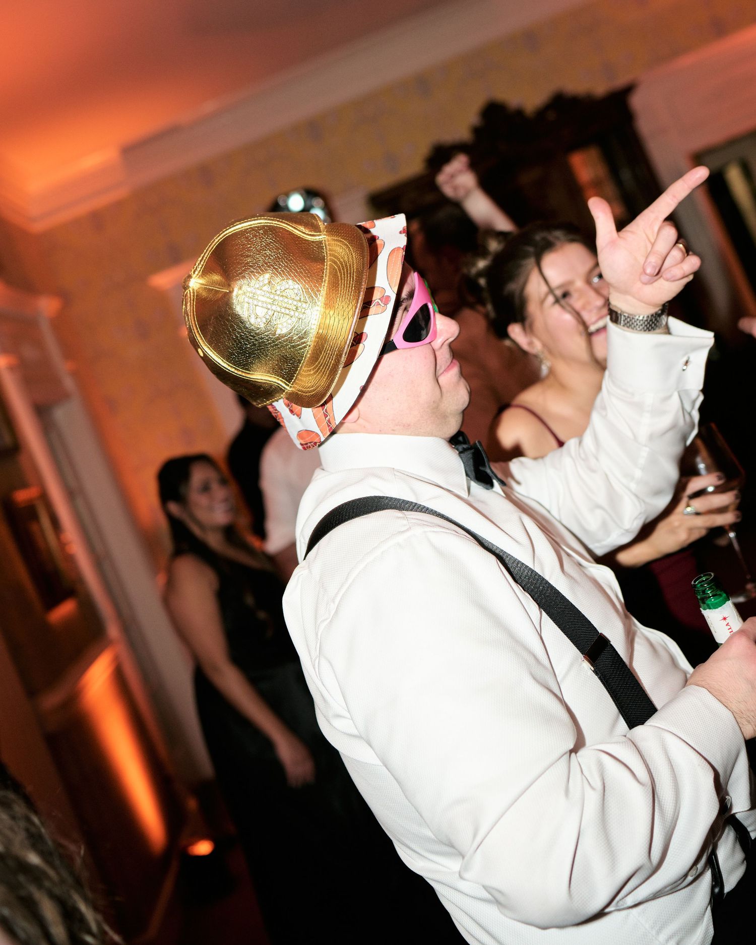 Person wearing gold hat and pink glasses dancing at a wedding reception party with guests in background.